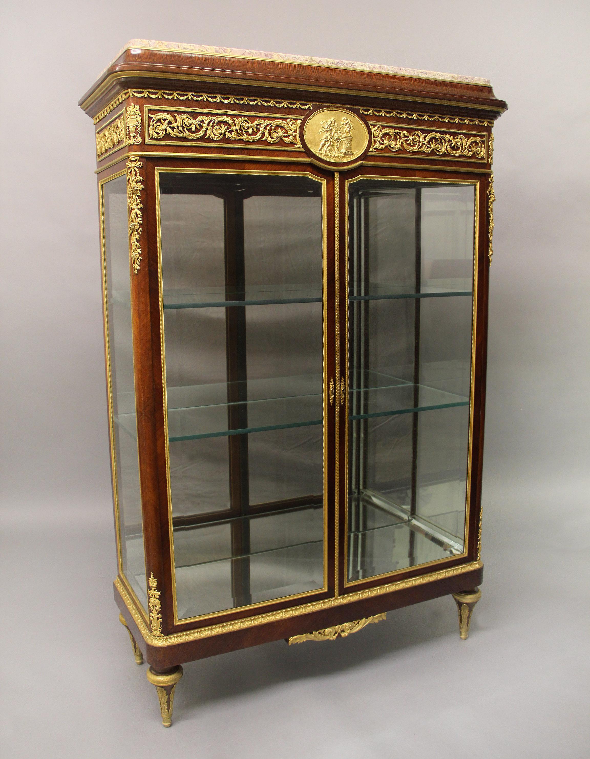 An exceptional quality late 19th century gilt bronze mounted Louis XVI Style Vitrine.

The marble top above ormolu frieze and scrolling foliage centered by an bronze oval plaque of two women and frolicking cherubs, the corners with flowering urns