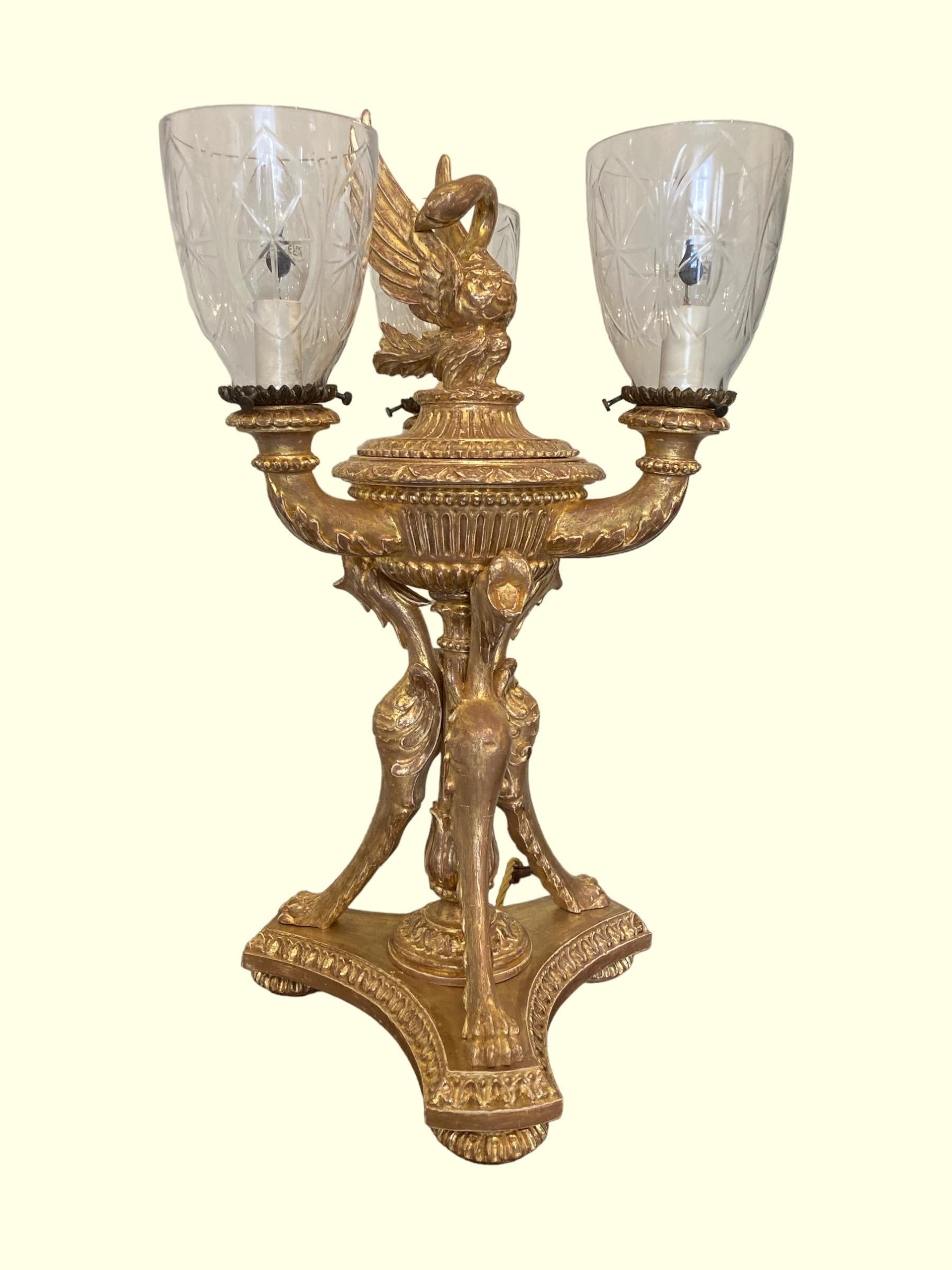 An extremely decorative Italian 19th-century giltwood three-light table lamp featuring a central swan surrounded by three giltwood candle arms with glass shades.

The swan is mounted on a central urn-shaped support adorned with three single-footed
