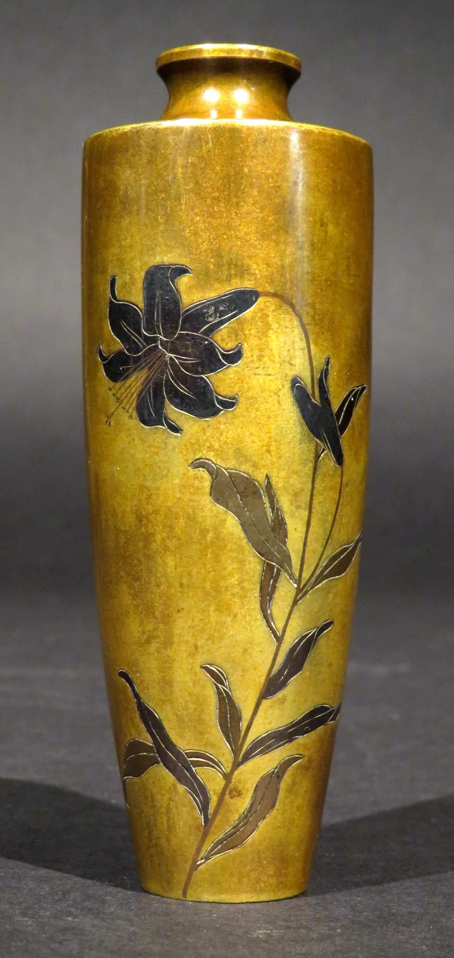 Il corpo cilindrico, piccolo e finemente proporzionato, illustra un motivo naturalistico di un fiore di giglio argentato che spunta da uno stelo frondoso, decorato con le tecniche Shibuichi e Shakudo; la parte inferiore della base è firmata in