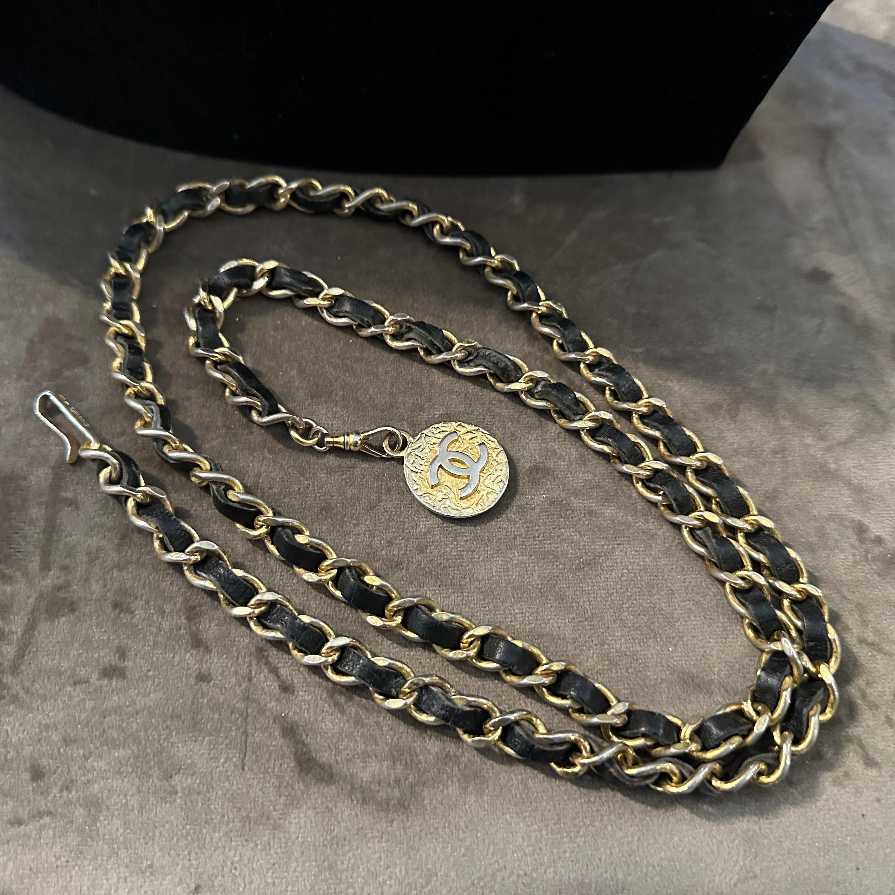 This image showcases an iconic Chanel leather and metal chain necklace, a hallmark of the brand’s timeless and luxurious aesthetic.  The necklace features Chanel’s signature interwoven chain, where black leather is intricately threaded through