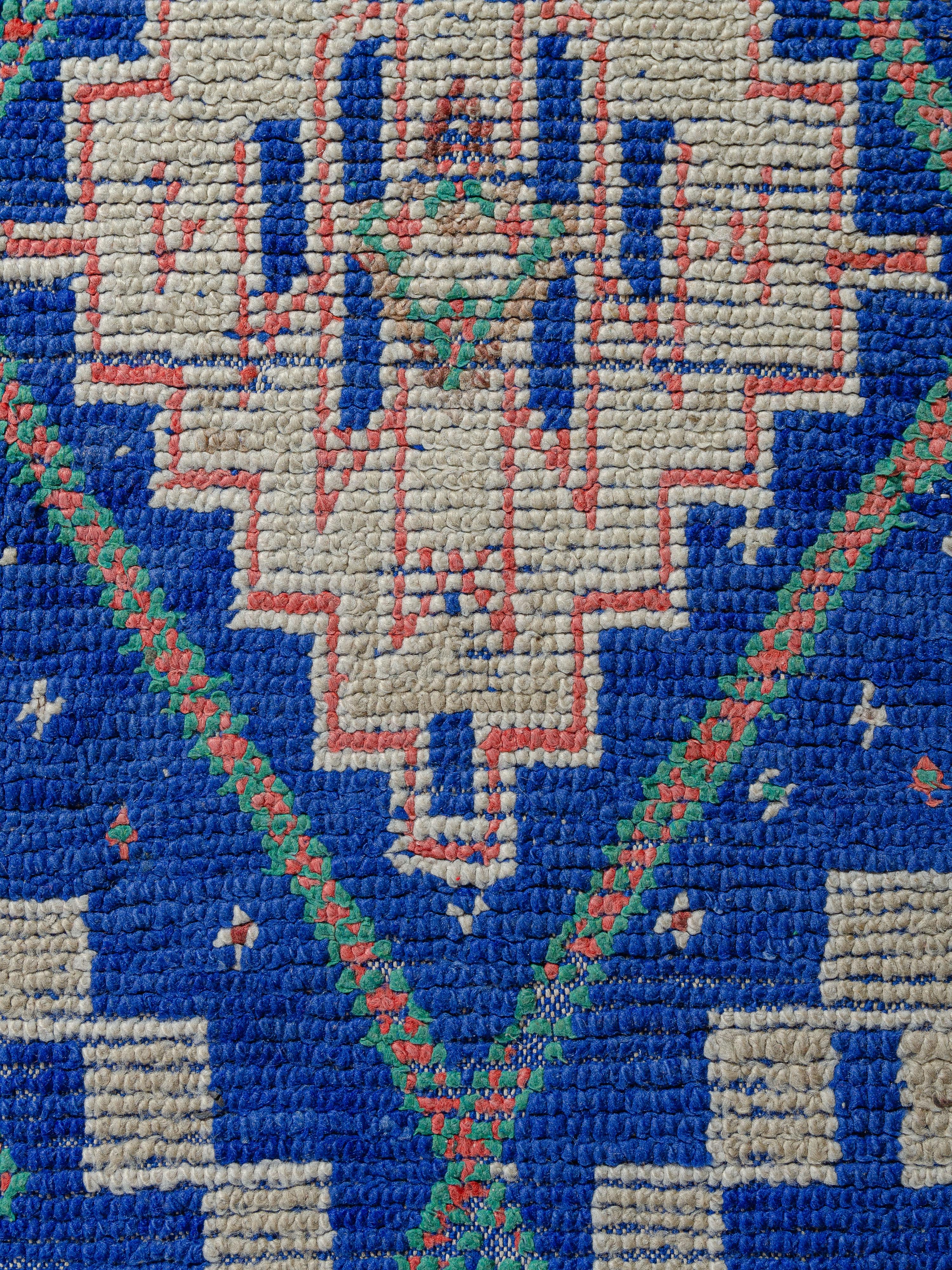 A small yet impactful vintage Beni M’Guild example featuring a stunning cobalt blue ground. The bold graphic motifs surrounding the central lozenge network are knotted in contrasting mint green, coral red, and ivory. Intricate side borders are