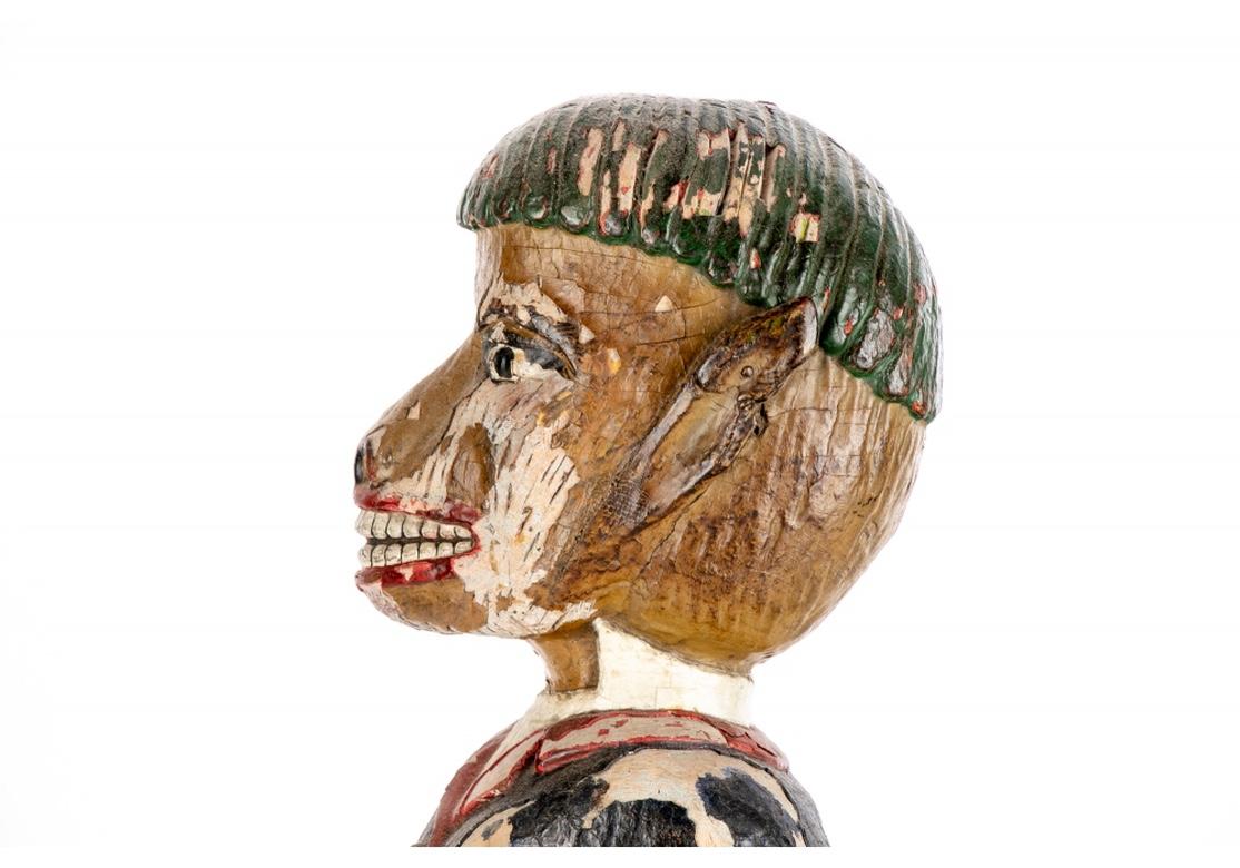 A hollow finely carved wood macaque figure dressed as a human with short green hair, a blue jacket and yellow slacks. He stands and holds a walking stick in his left hand and crosses his right hand over his chest. His red lips smile as he looks
