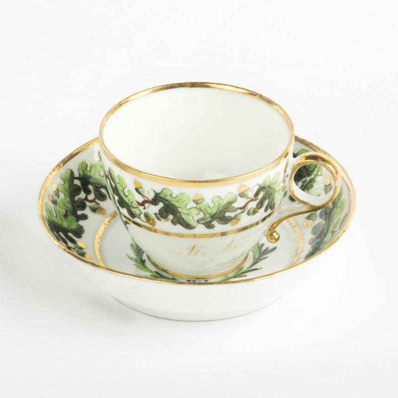Both pieces are delicately painted in underglaze blues and greens with over glaze hand gilding.  Each has a border of oak leaves and gilded acorns, a central fouled anchor within a victor’s laurel wreath and gilt borders.  The gilt inscriptions