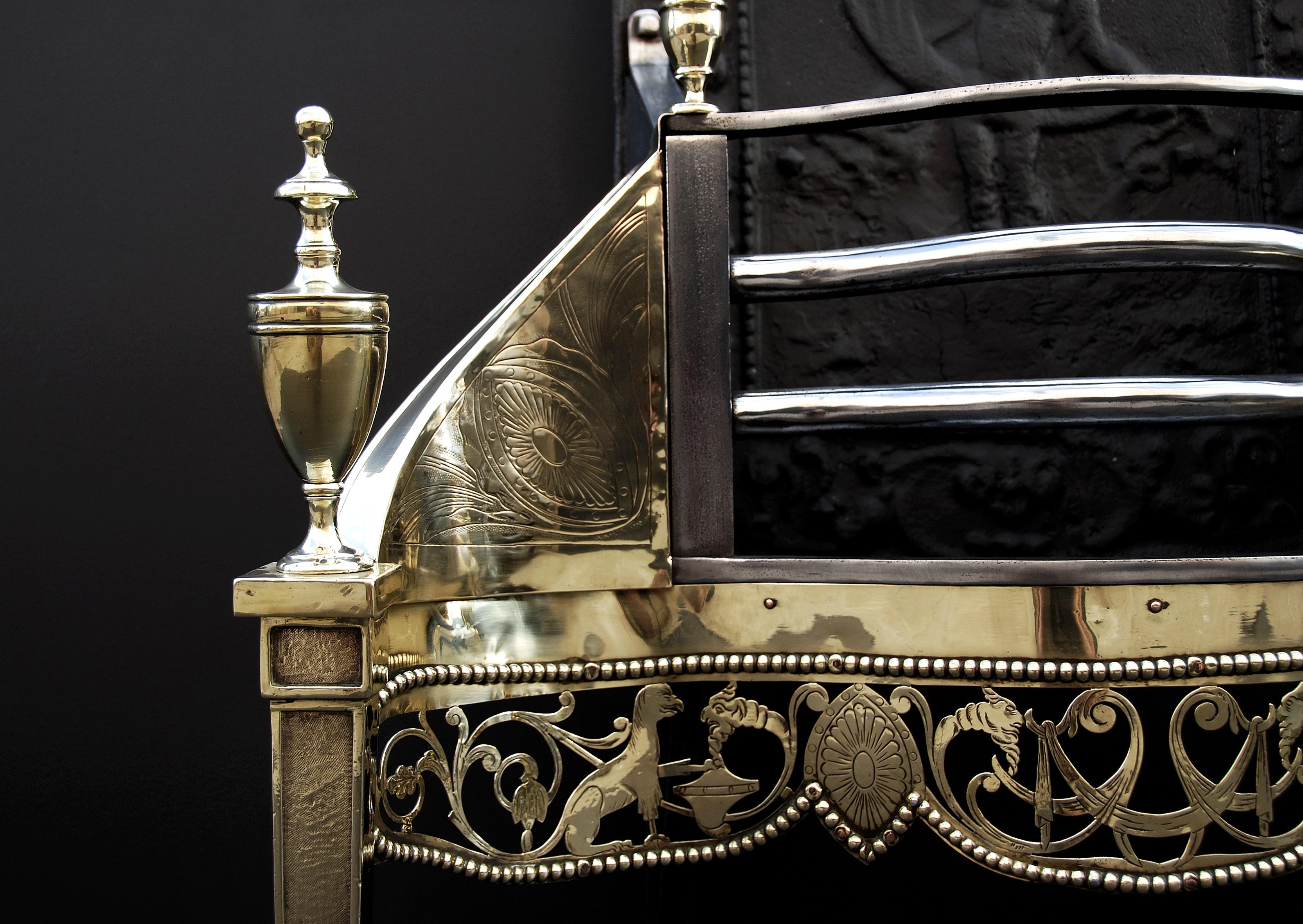 An imposing Georgian style brass firegrate. The pierced brass fret engraved with griffins and foliage with beading above and below, the tapering panelled legs surmounted by large brass urn finials, the shaped iron front bars flanked by engraved side