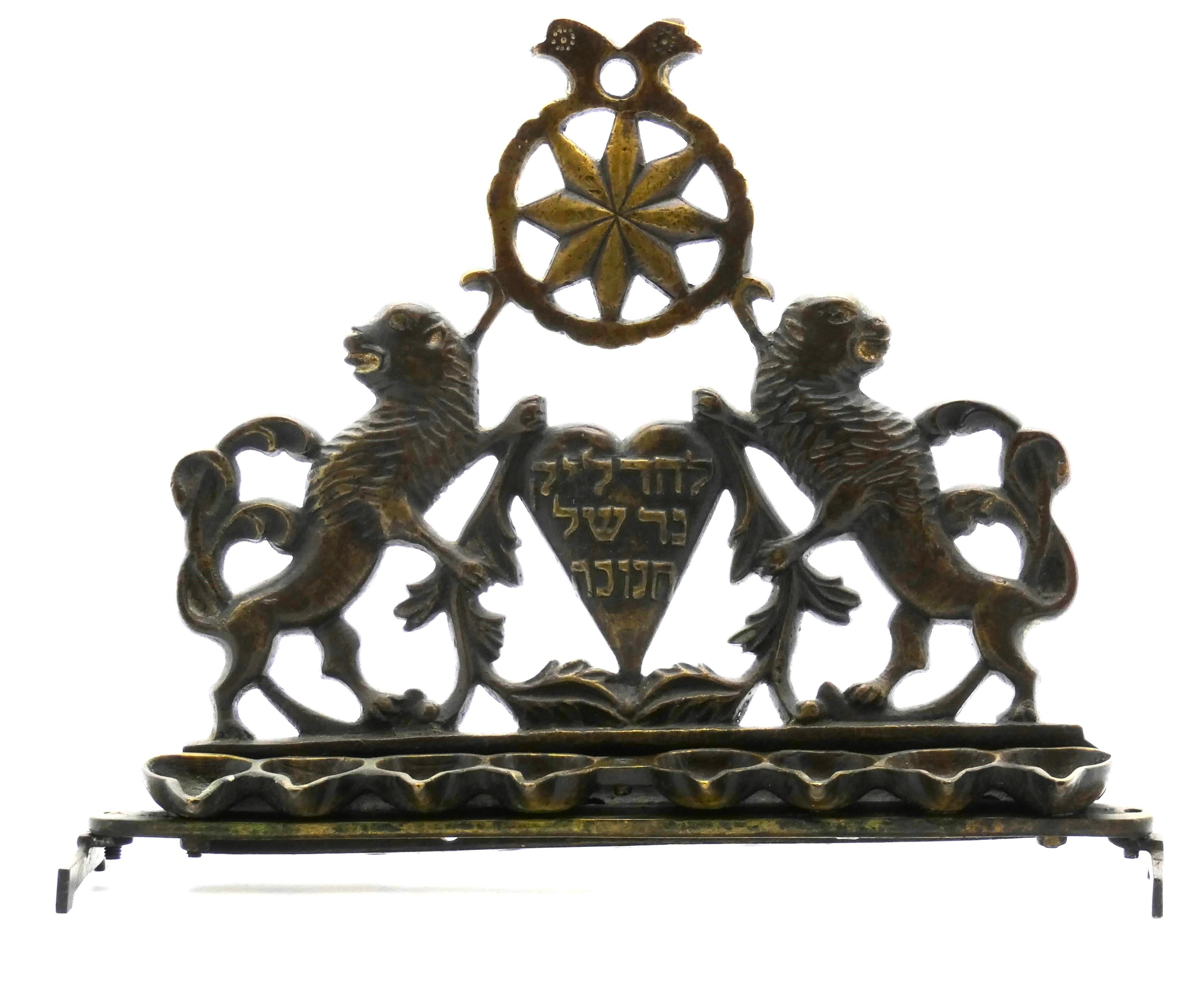 This spectacular Hanukkah lamp is ornately fashioned with a cast backplate featuring a heart shape at center flanked by two rampant Lions of Judah fiercely facing away from one another clutching flowing foliate branches. 

These magnificent lions