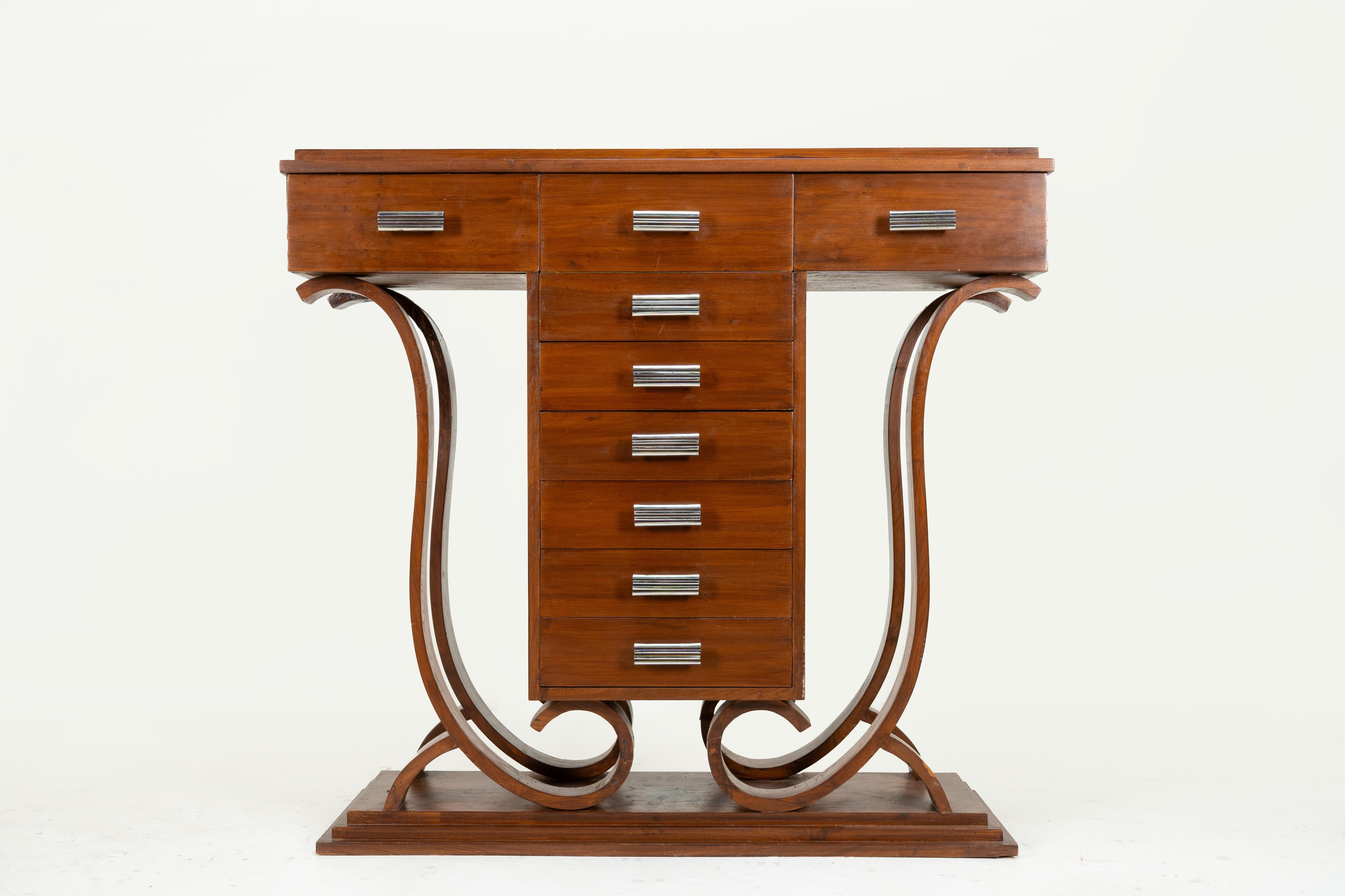 An exceptional Indian Art Deco console is from Ahmedabad, c.1930, masterfully crafted from teak. Its defining feature is the pair of scrolled legs, rising in sweeping curves from the solid plinth base. The bold vertical column of drawers provides