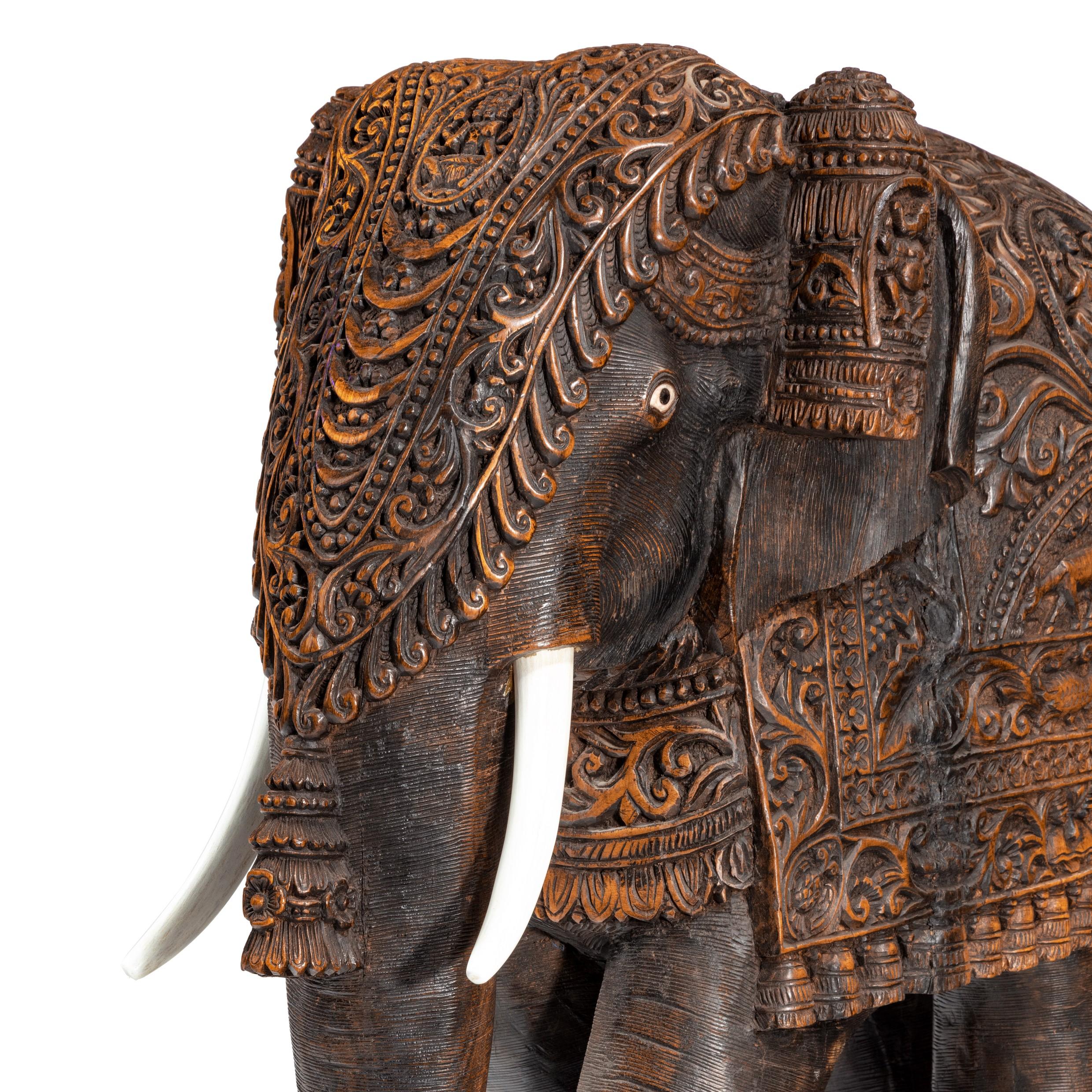This hardwood stool is in the form of a caparisoned Indian elephant with wooden tusks, shown walking forwards with a curled trunk, the ceremonial trappings comprising a triangular head piece tapering to a large tassel and knee rolls beside the ears,