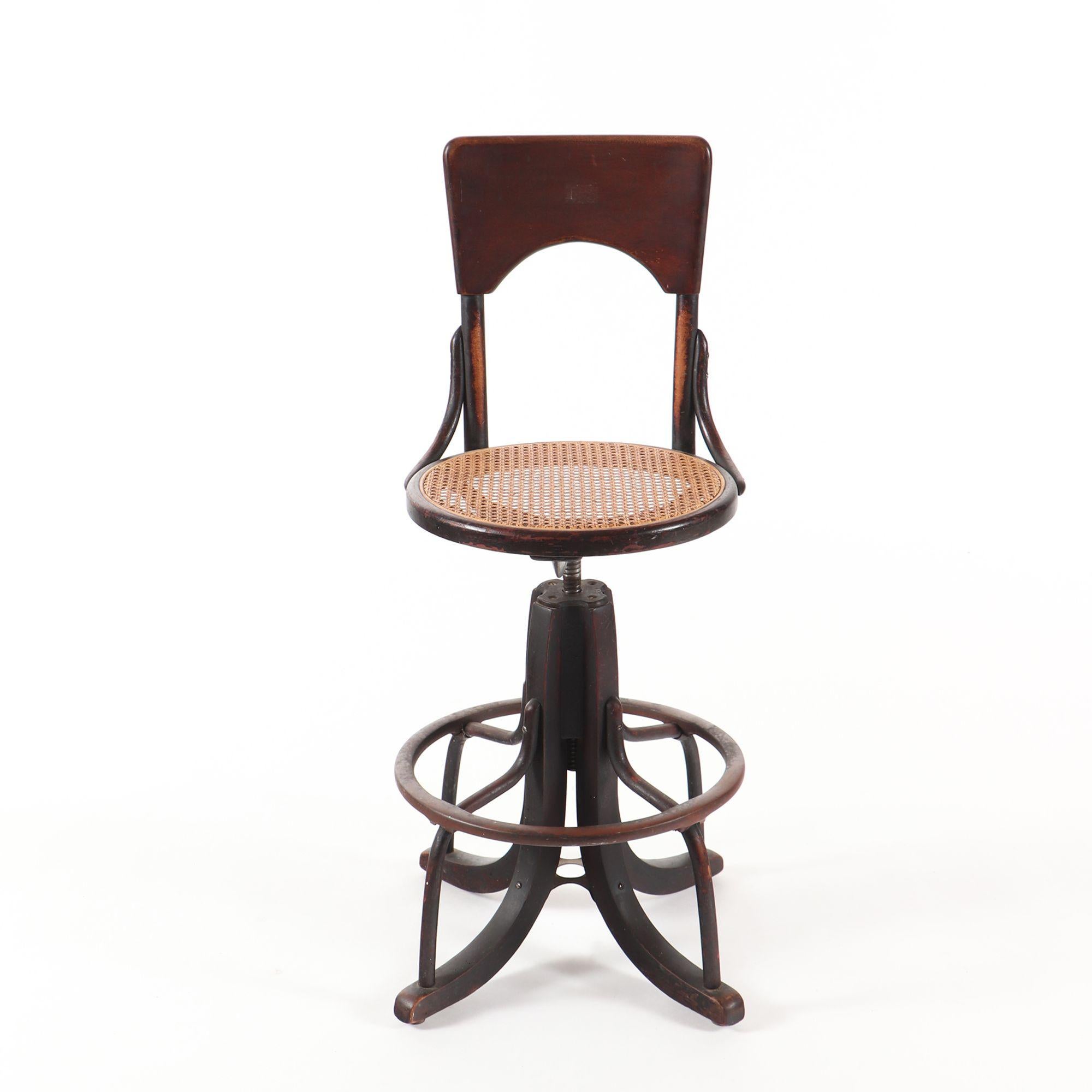 An Industrial swivel stool or telephone operator's chair, circa 1930.
Having a tall wood back with cane seat, resting on downswept legs with circular foot rest.