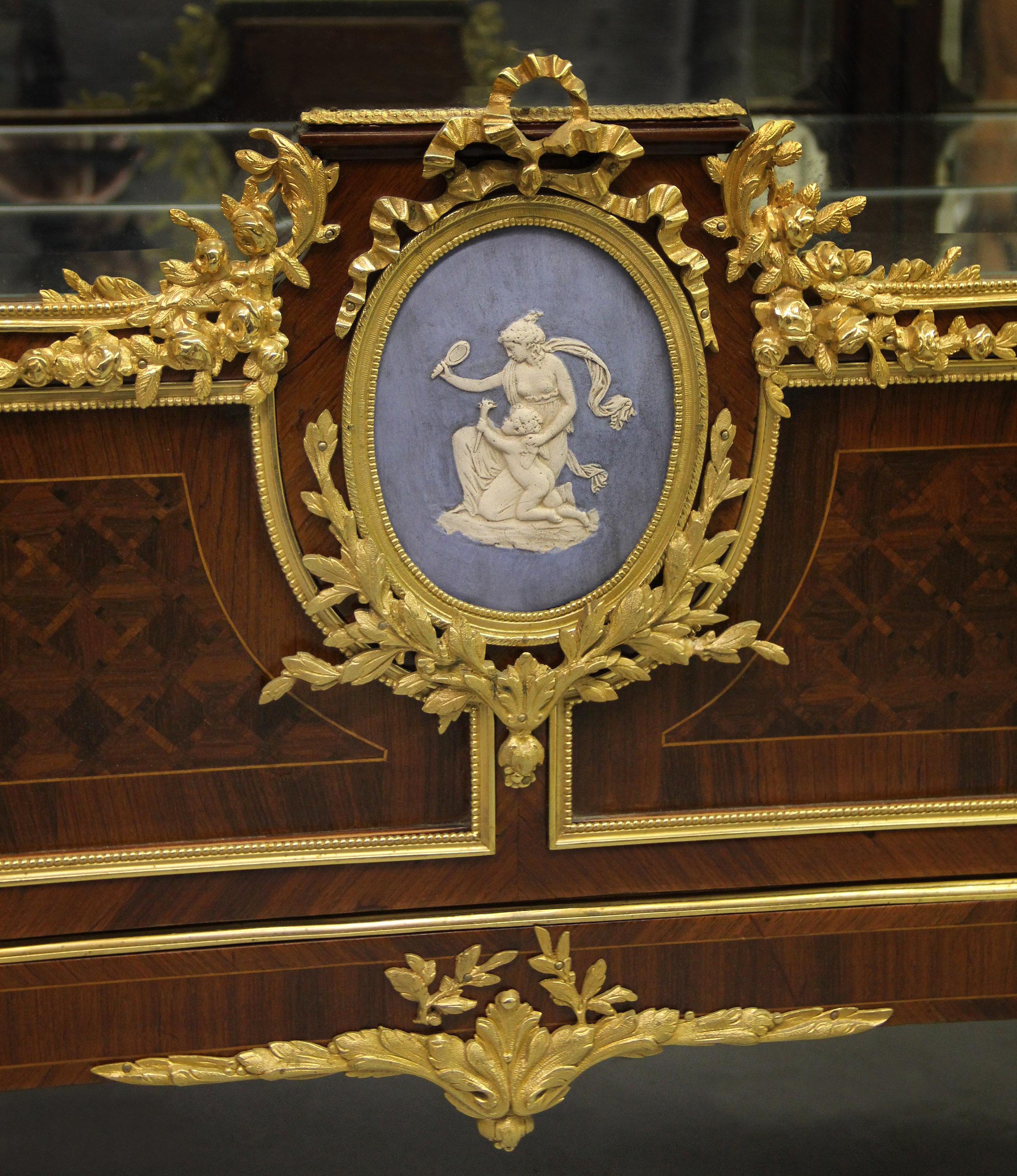 An interesting late 19th century Louis XVI style gilt bronze and wedgwood Mounted parquetry vitrine

The breakfront marble top above a scrolled frieze cast with motif depicting a pair of rams centered by cornucopia and a maiden, over a single