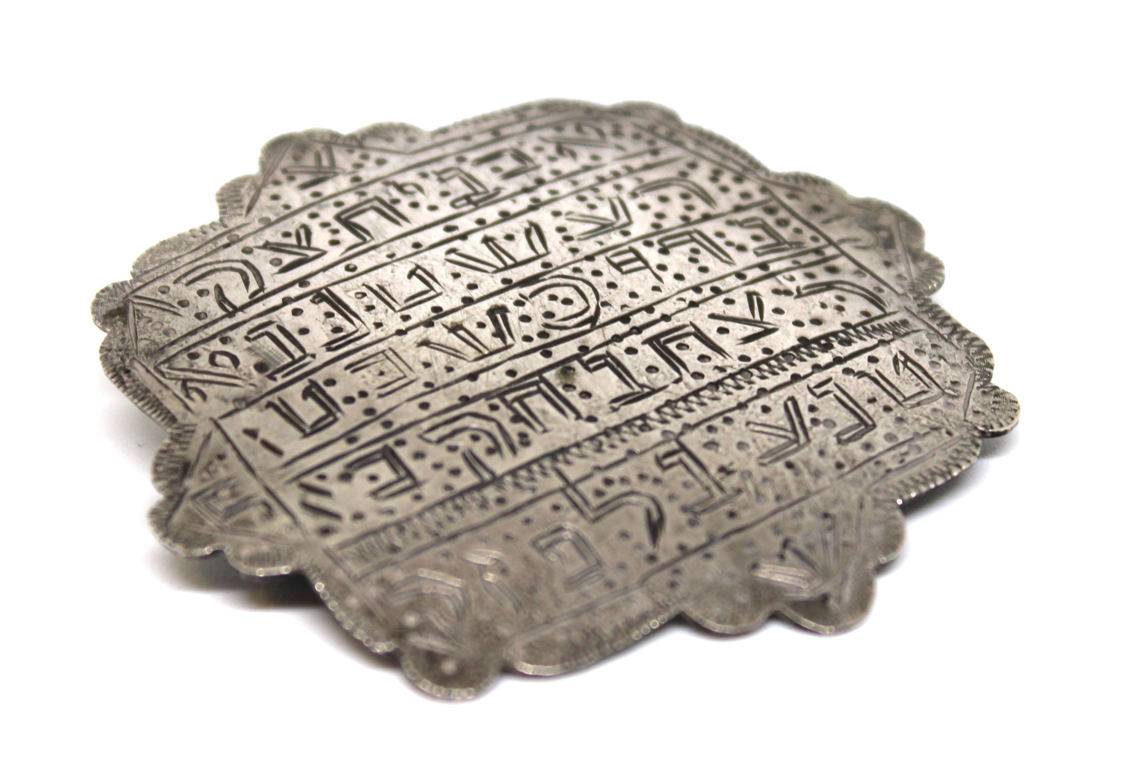 Cette magnifique amulette ancienne en argent provenant d'Iran est un bel exemple de l'artisanat juif persan. Sa forme festonnée, semblable à celle d'un bouclier, est méticuleusement gravée à la main d'inscriptions hébraïques denses disposées en
