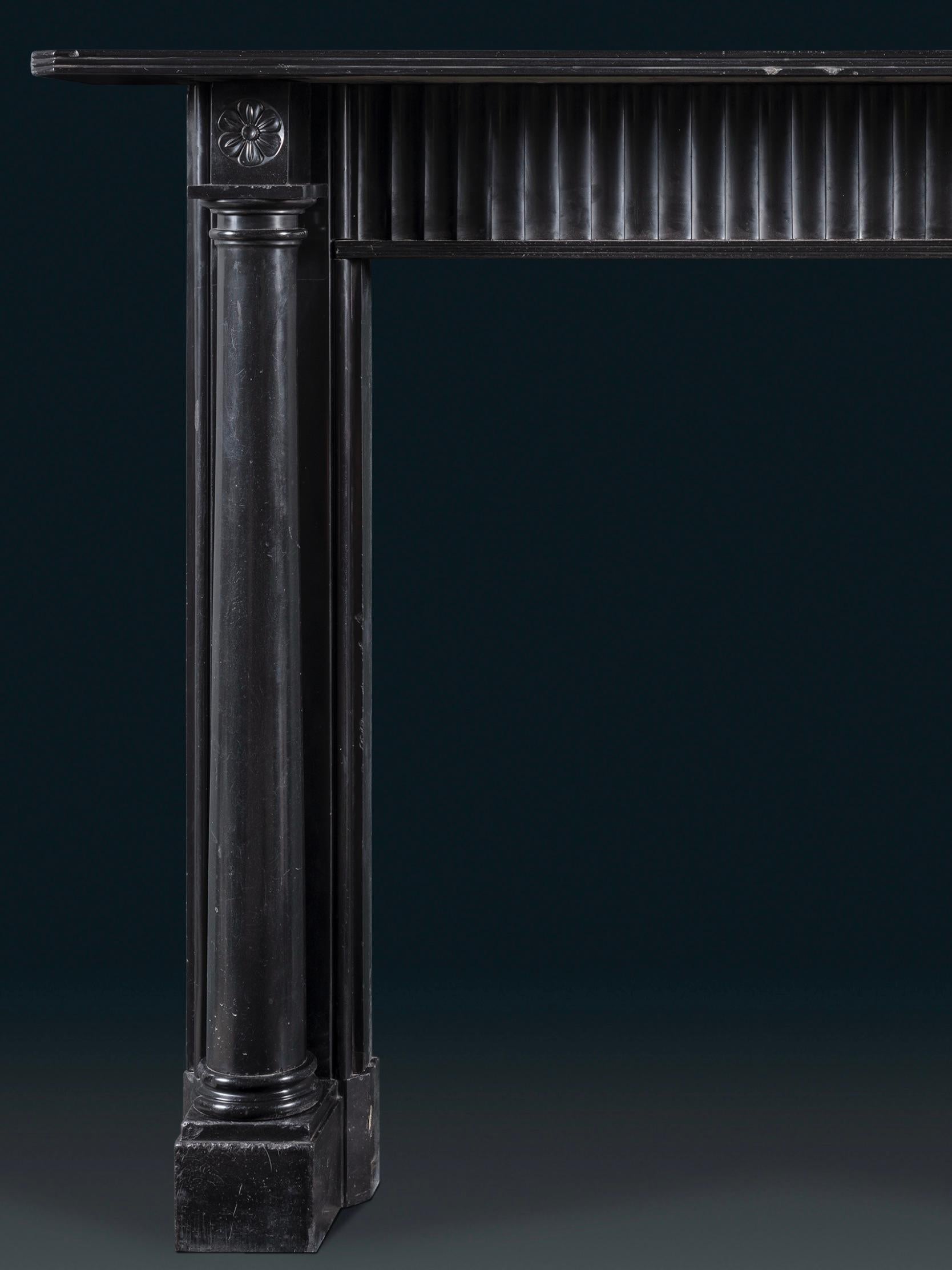 Ein aus dem Vollen geschnitzter Regency-Kamin, dessen Pfosten die Form von freistehenden dorischen Säulen auf Sockeln haben, die auf quadratischen Fußblöcken ruhen. Der Fries mit geriffeltem Motiv trägt ein einfaches, profiliertes Regal. Auf den