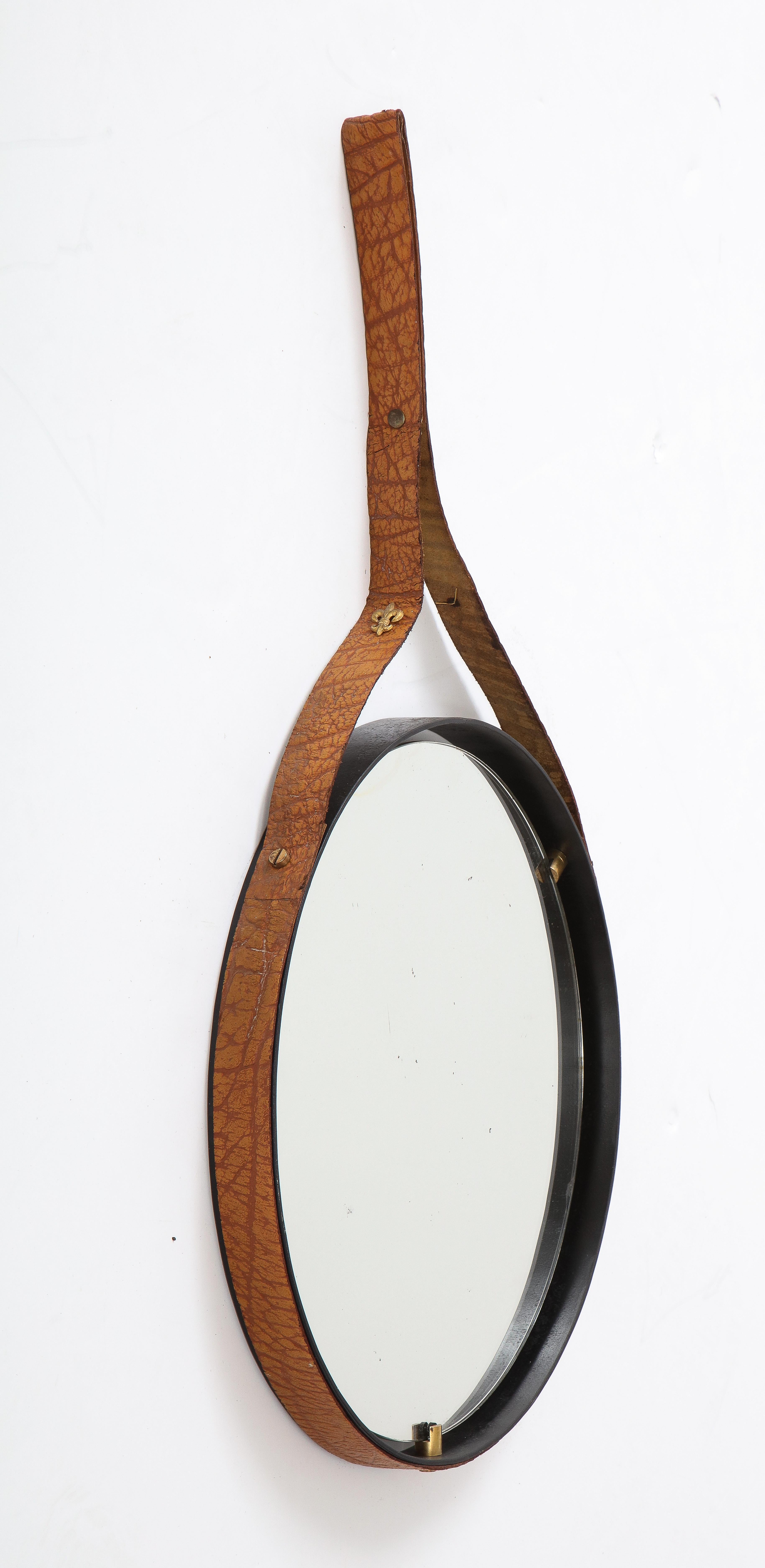 An Italian 1960's circular floating mirror, with round black iron frame which holds the floating mirror secured with brass pegs. The iron frame is wrapped with a textured brown leather with a hanging strap with brass fleur de lis detailing. A