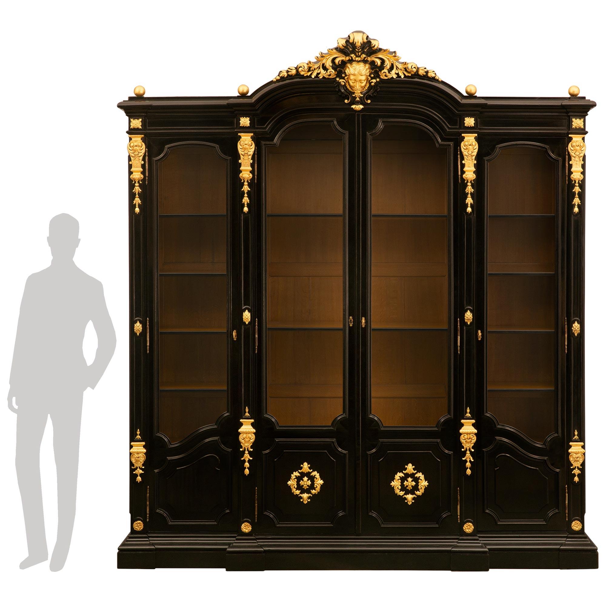 A most impressive and large scaled Italian 19th century Rococo Revival st. Ebonized Fruitwood and giltwood cabinet/vitrine. The four door cabinet is raised by a mottled plinth below the two central doors showcasing mottled square panels with concave