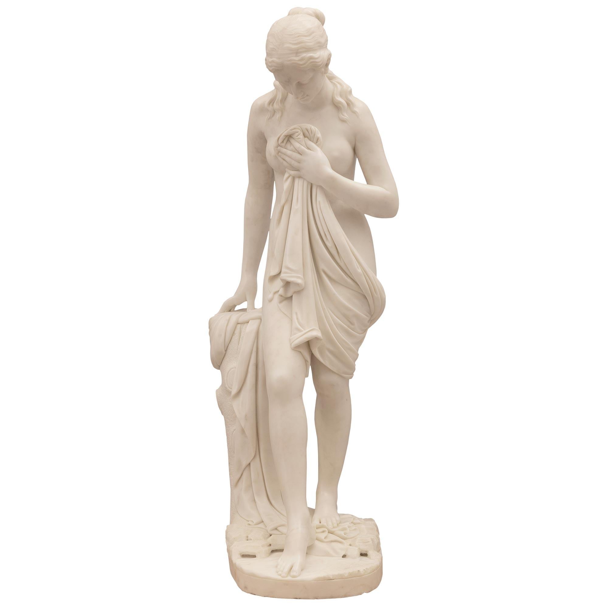 A stunning and extremely high quality Italian 19th century white Carrara marble statue of a beautiful bathing woman. The statue is raised by a ground designed base where the richly sculpted maiden is standing next to a tree trunk. She is draped in a