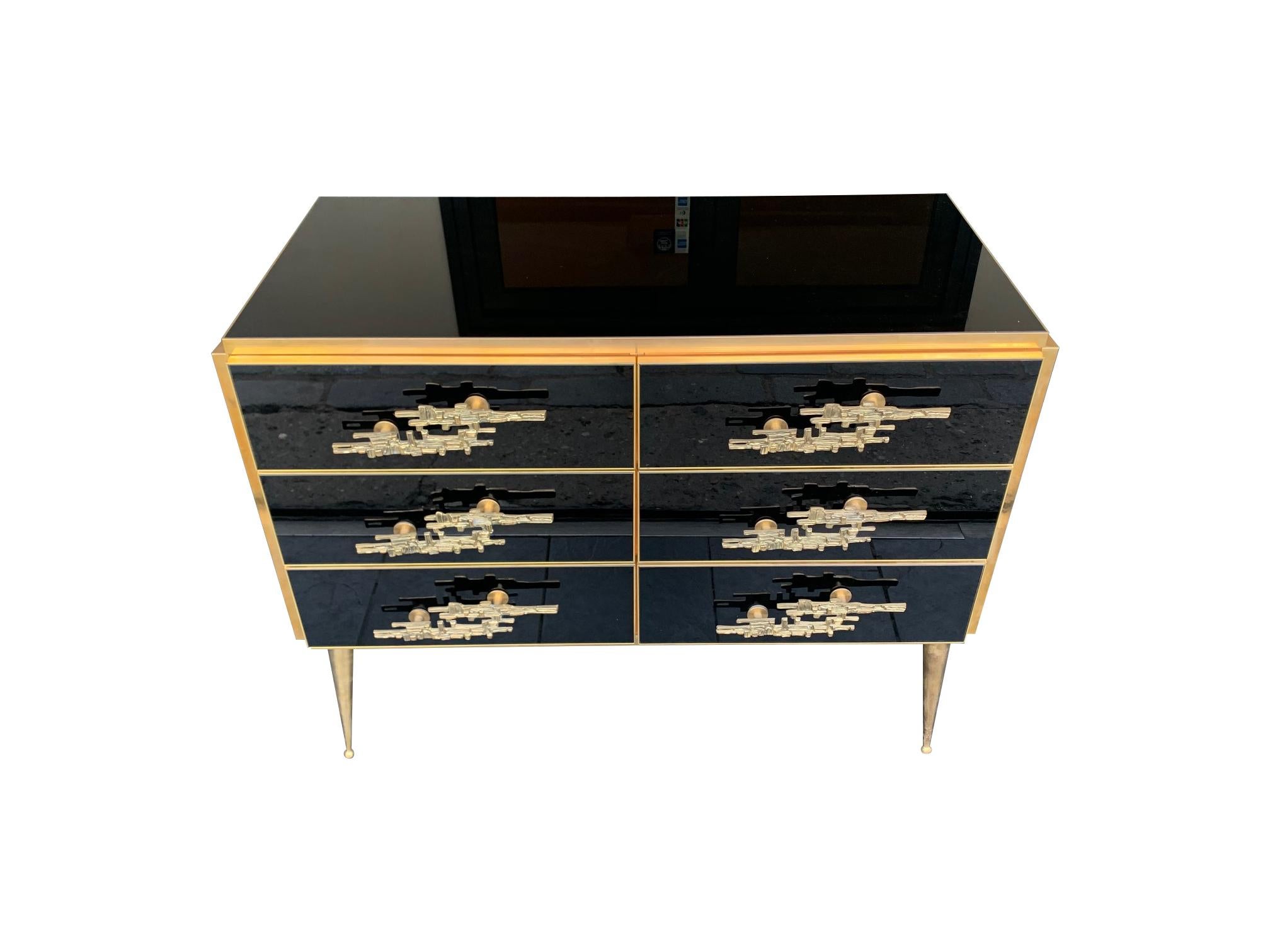 An Italian black glass and solid brass chest of drawers with six drawers with a solid wood interior and stunning Brutalist brass handles. The chest is mounted on 1950s style pointed brass legs. 

This piece in the listing is available to buy now,