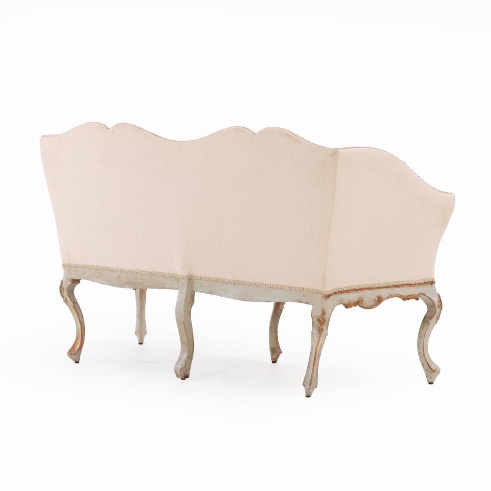 An elegant 18th century Italian settee, finely carved and painted in soft tones, reflecting the restrained opulence of late Baroque or early Neoclassical design. The shaped frame features delicate hand-carved detailing along the apron and cabriole