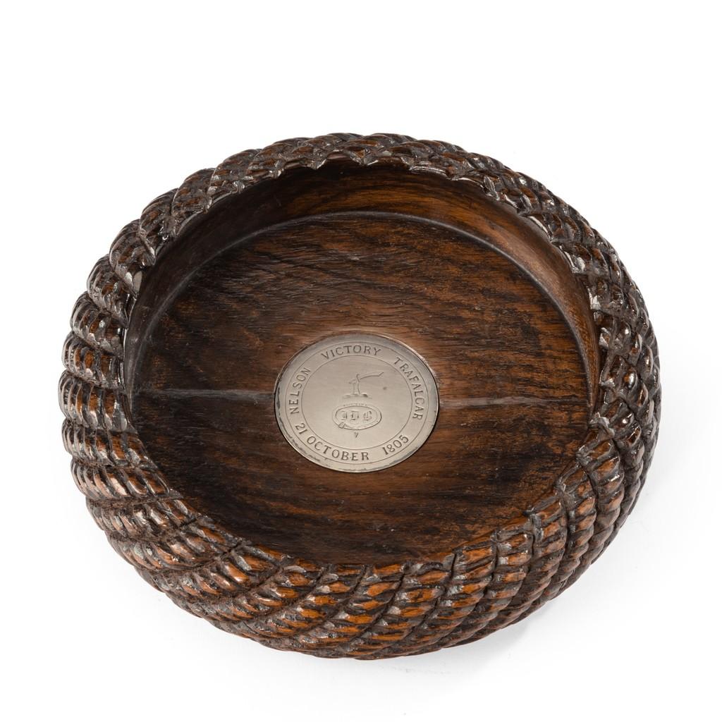 An oak wine coaster made from Victory timber, of circular form with carved rope twist sides enclosing a solid base set with a silver roundel inscribed ‘Nelson Victory Trafalgar 21 October 1805’ encircling a crest of a raised arm and cutlass and
