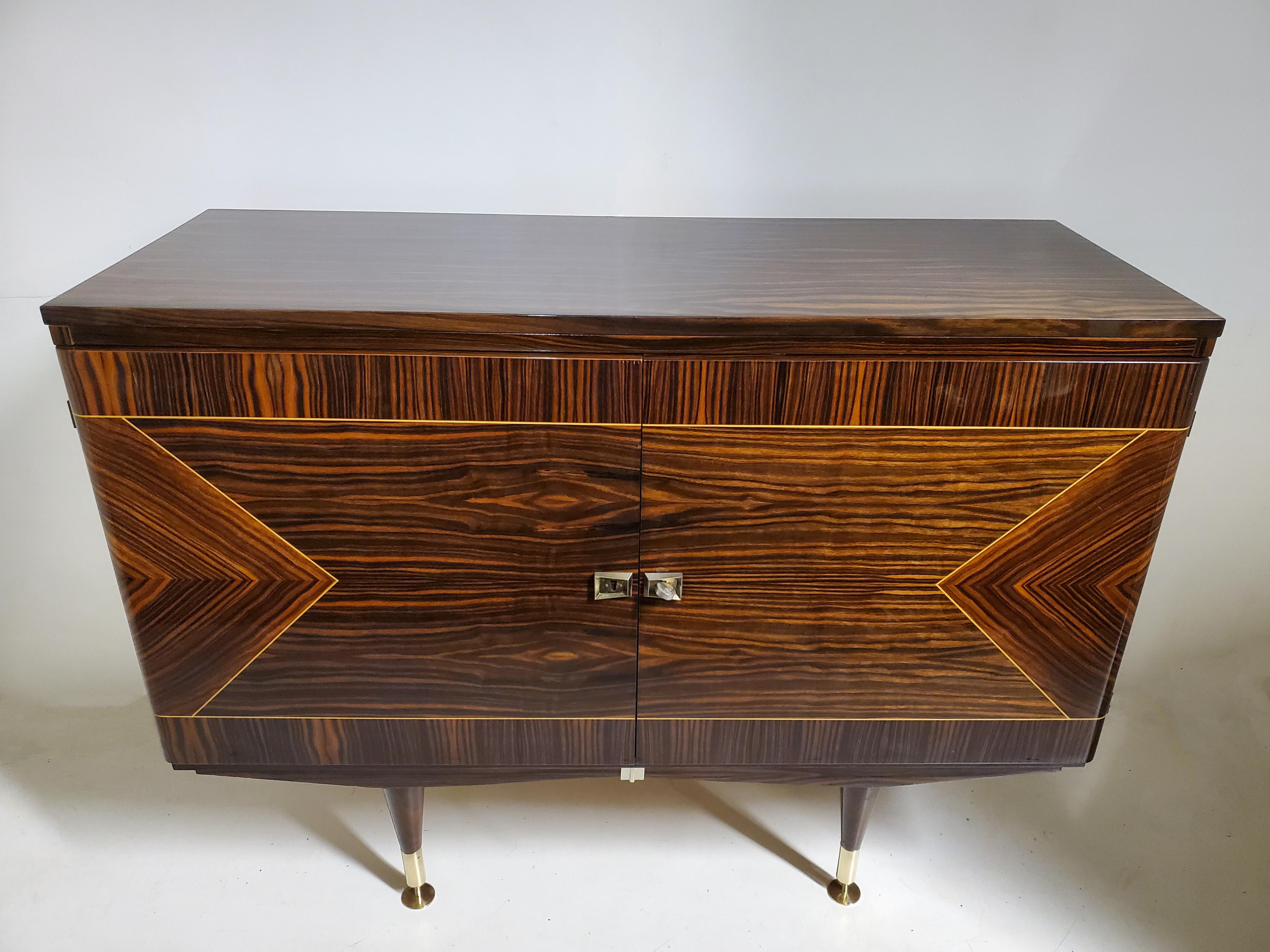 A petite scaled, macassar ebony vintage cabinet with double doors, featuring parquetry inlaid triangular, arrow like patterns flanking the sides of each door. 
The cabinet, angular and cubist in feeling is raised on tapered legs ending with brass