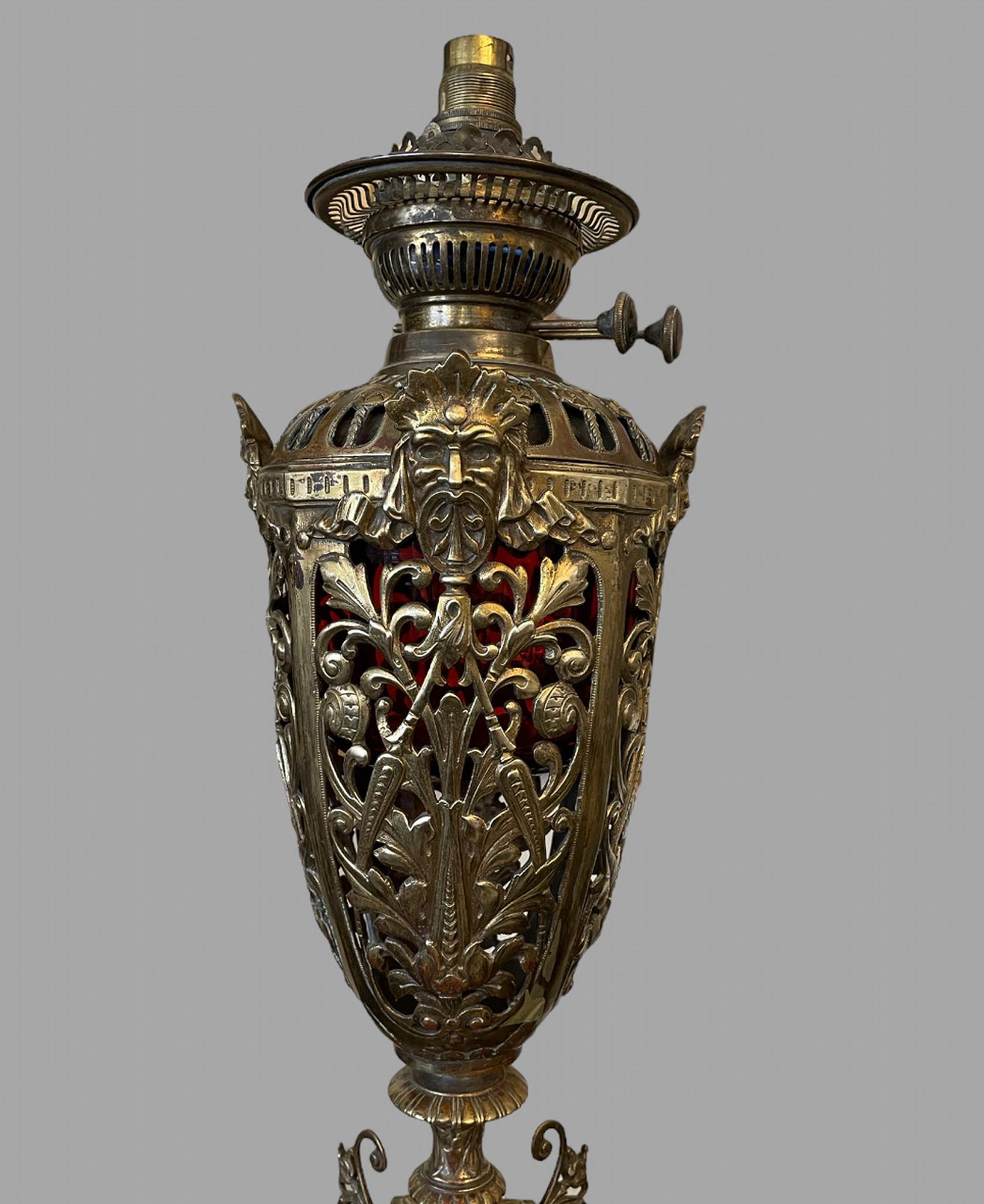 An Ornate Brass and Ormulu Oil Lamp with original reservoir. It has central masks on the square base of the deity Cernounnos who represented prosperity. The upper vase shaped part of the lamp is beautifully wrought and pierced with leaves and