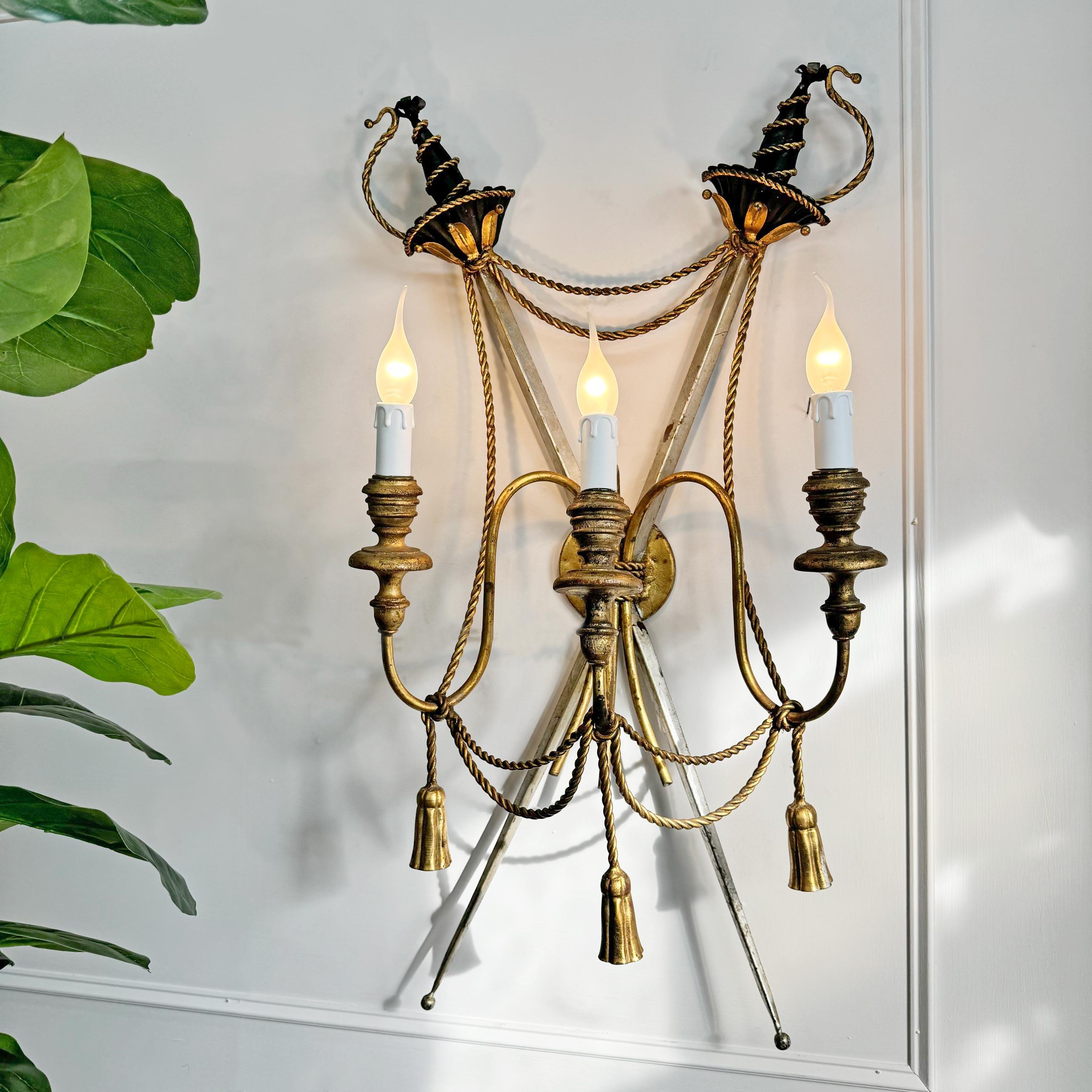 An extremely rare and incredibly ornate Italian wall light, dating to the 1950's, in the empire style and modeled as a pair of crossed swords, in gilt and silver gilt, decorated with gold rope twist and tassel to the front, the three lamp holders in