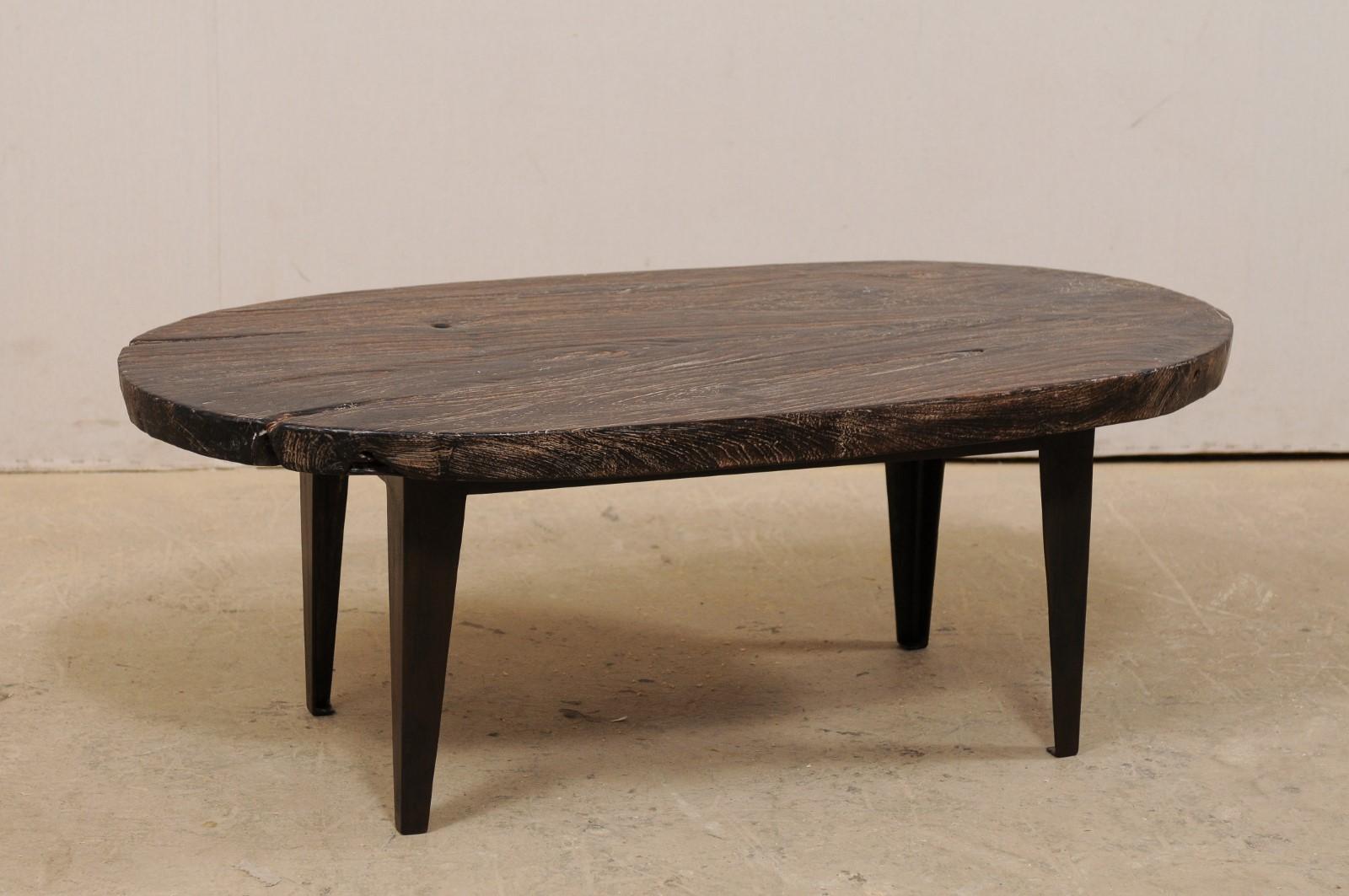 A custom created teak wood top coffee table with iron base. This rustic yet chic coffee table has been fashioned from an oval-shaped piece of teak wood slab, which has been presented upon four custom iron legs. The wood has been carbonized, which is