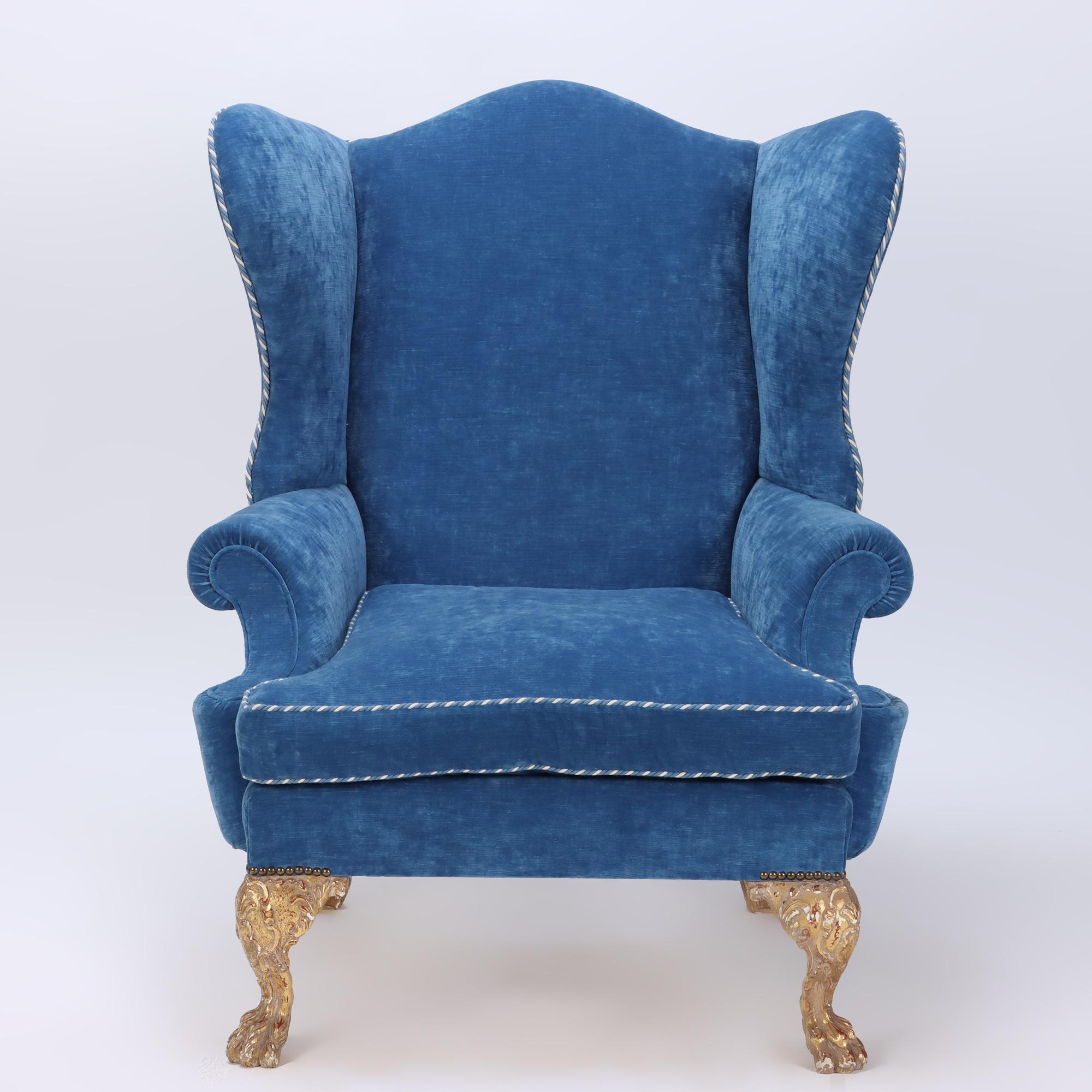 An oversized and exaggerated blue upholstered wing chair and ottoman C 1900 with gilt carved feet. A statement piece for sure!. 
Ottoman: 25