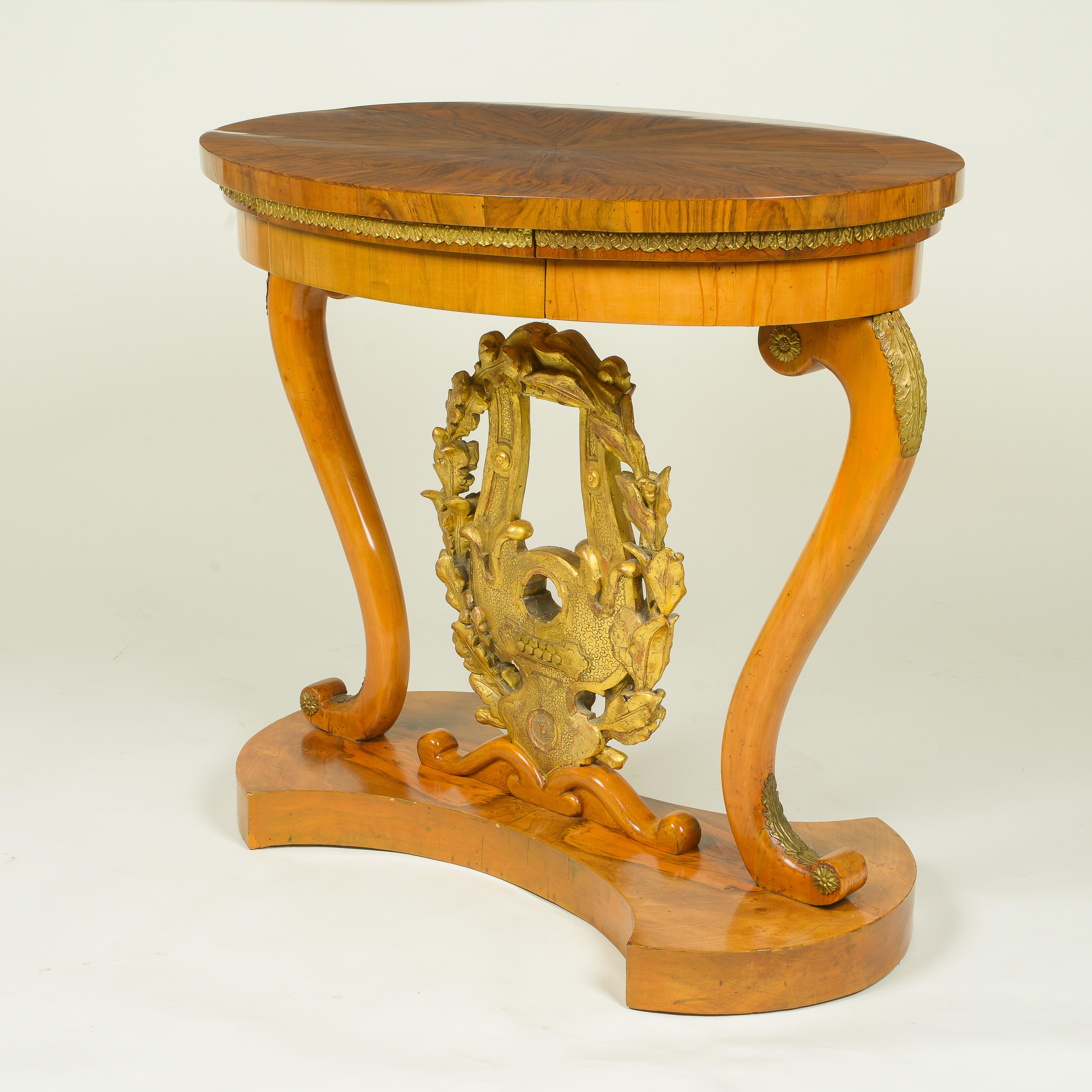 The oval top veneered in a radiating pattern with highly figured walnut, with stiff-leaf gilt-metal banding, over a frieze drawer; raised on gilt-metal mounted fruitwood volutes and a carved giltwood lyre encircled by a laurel wreath, both emblems