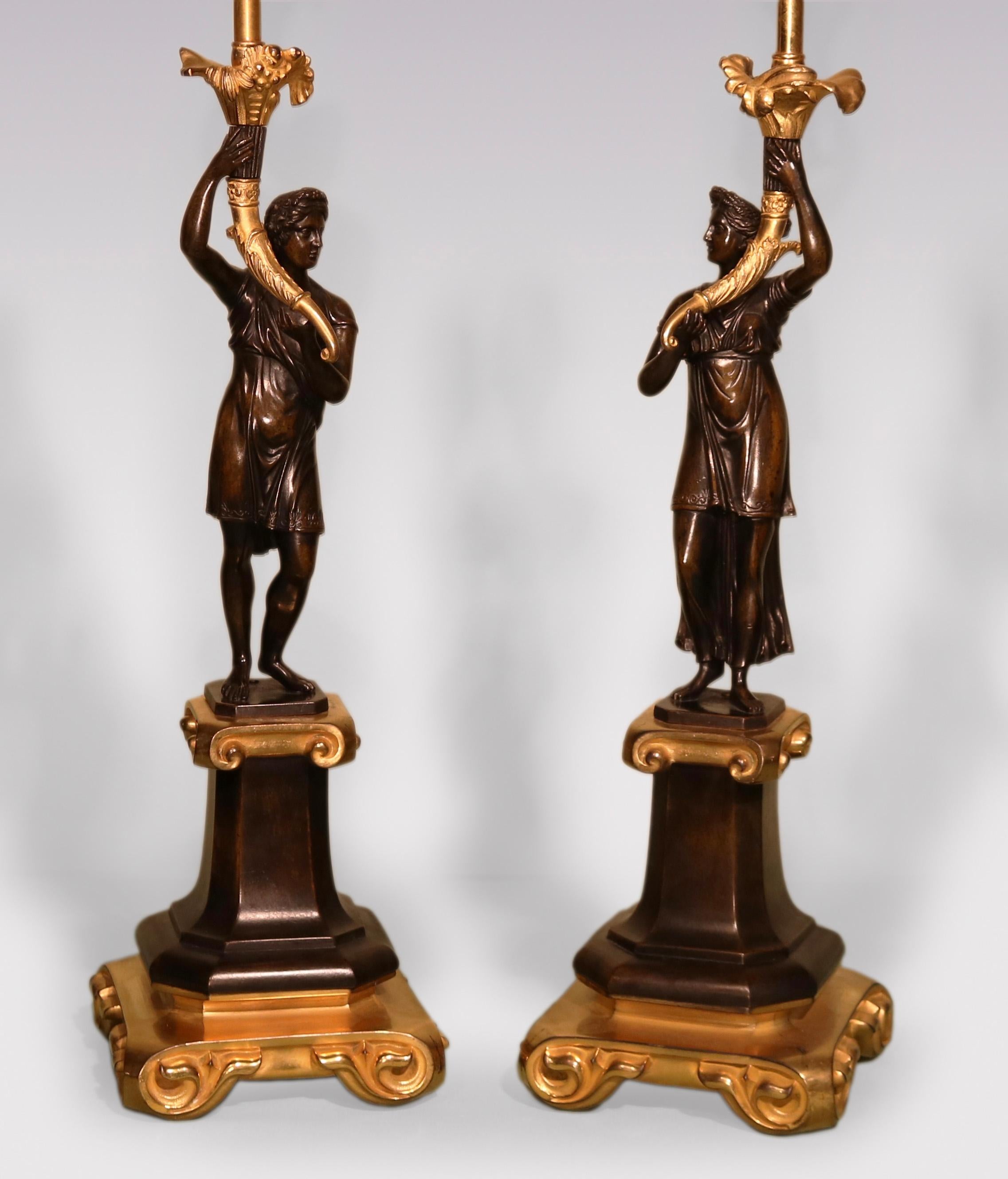 An unusual pair of early 19th century Regency period Bronze and Ormolu Candlesticks with male and female classical figures holding ‘Cornucopia sconces’ standing on scrolled platforms, raised on shaped square plinths ending on bold scrolled feet.