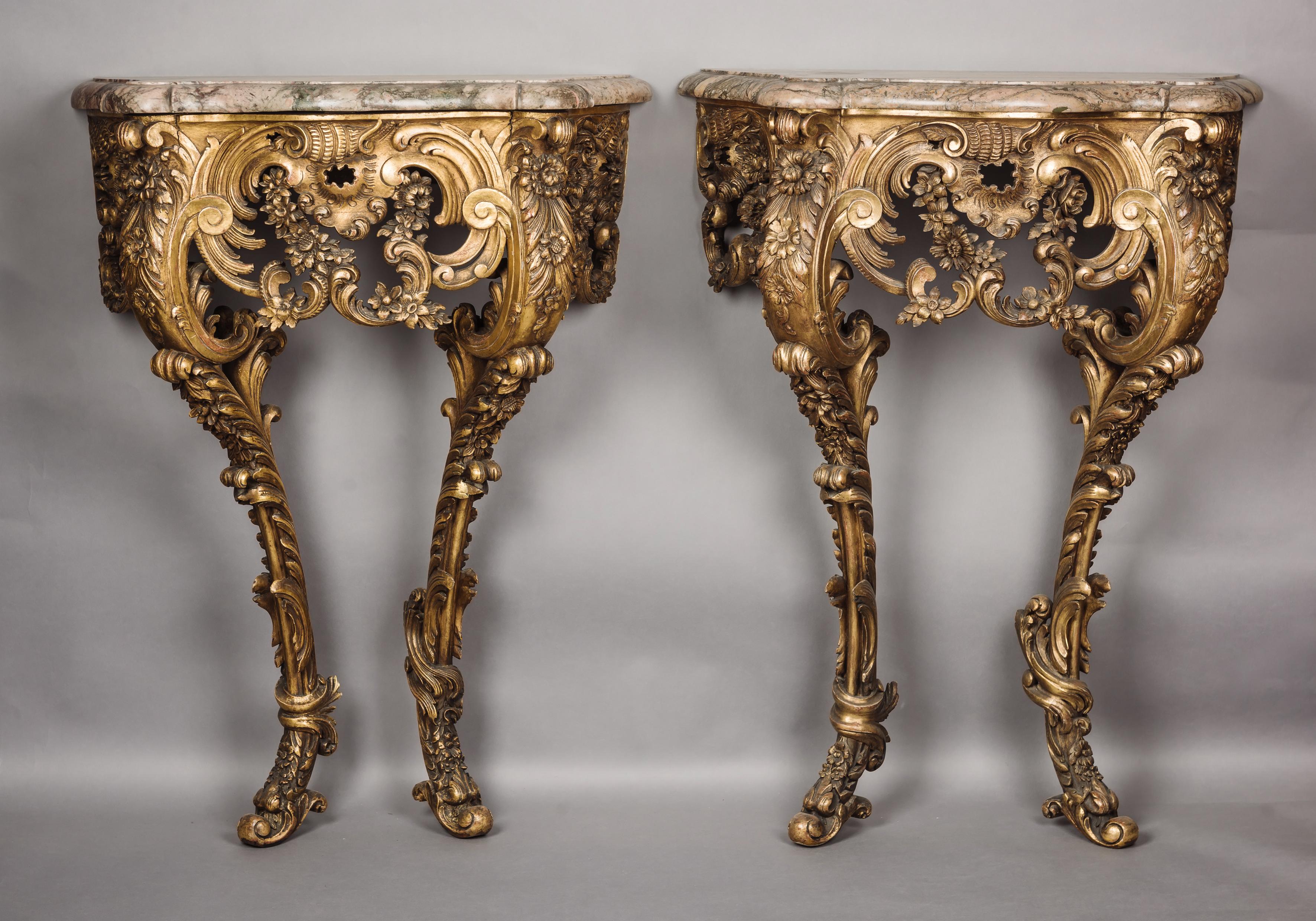 An Unusual Pair of Petit Rococo Revival Giltwood Console Tables. 

French, Circa 1860. 

The console tables each have a moulded marble top of serpentine outline above a conforming giltwood frieze elaborately carved in the rococo style with