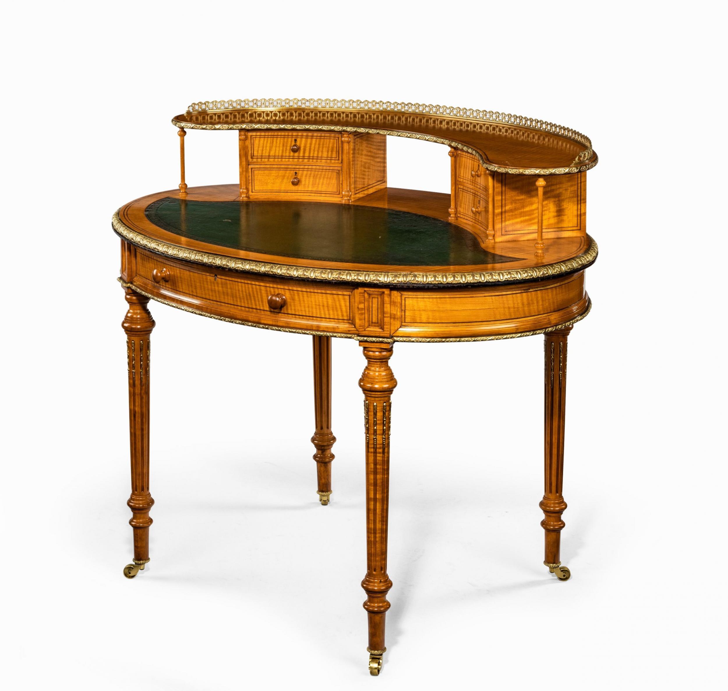 An unusual Victorian freestanding oval satinwood desk, the leather-inset top supporting a shaped open shelf with an ormolu gallery above two pairs of small drawers with a bookshelf between, flanked by slender turned columns, the frieze with a