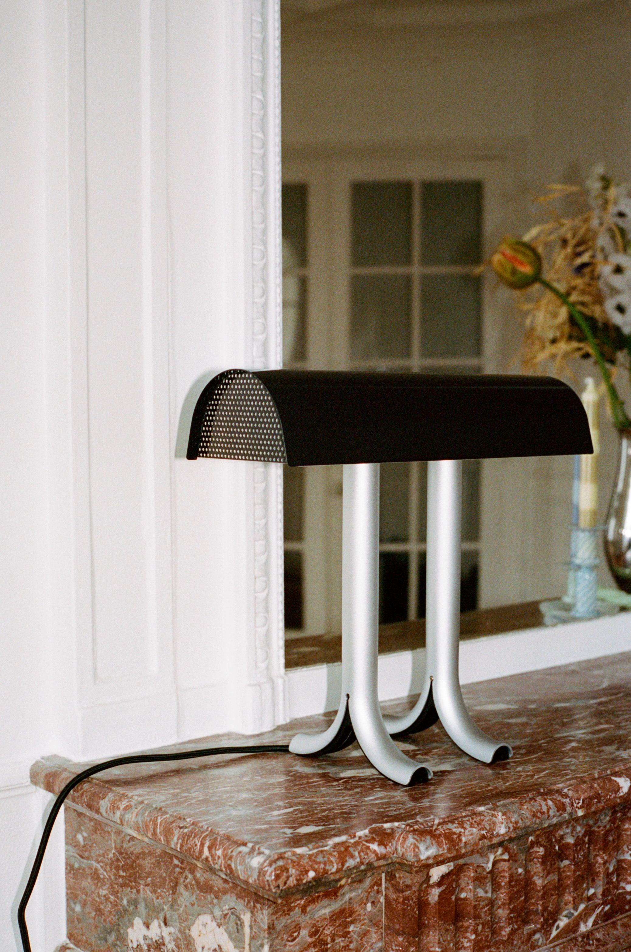 La lampe de table Anagram de Sam Weller allie fonctionnalité et attrait visuel pour créer une lampe de table à la construction robuste et à la personnalité forte. 
Doté d'un abat-jour brillant généreusement incurvé qui repose sur deux pieds fendus