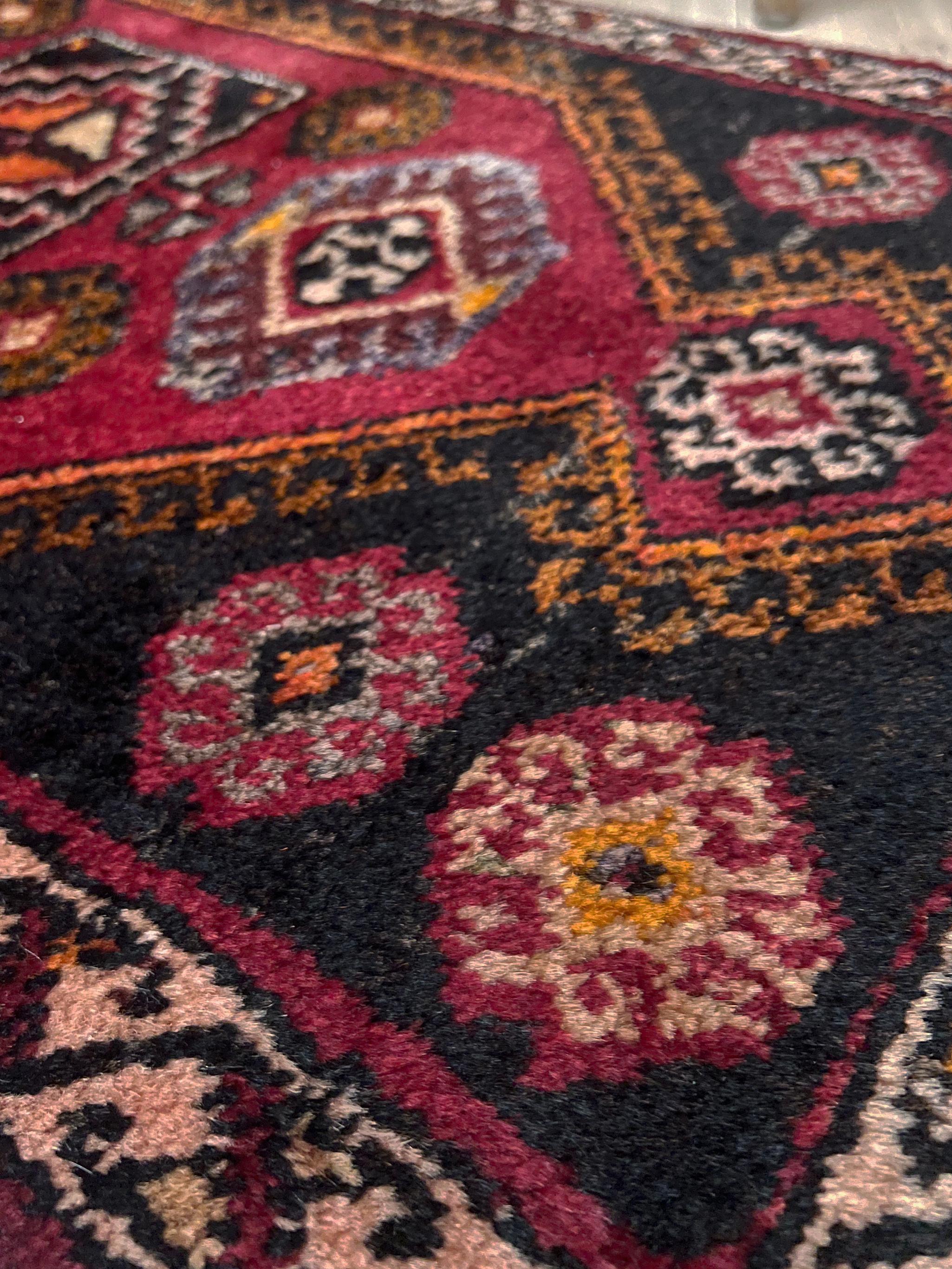 Un mini tapis turc vintage plein de caractère, tissé à la main avec de la laine douce filée à la main et présentant un emblème tribal central audacieux encadré par des tons rouge rubis, charbon de bois, ocre chaud et noyer terreux. Son format