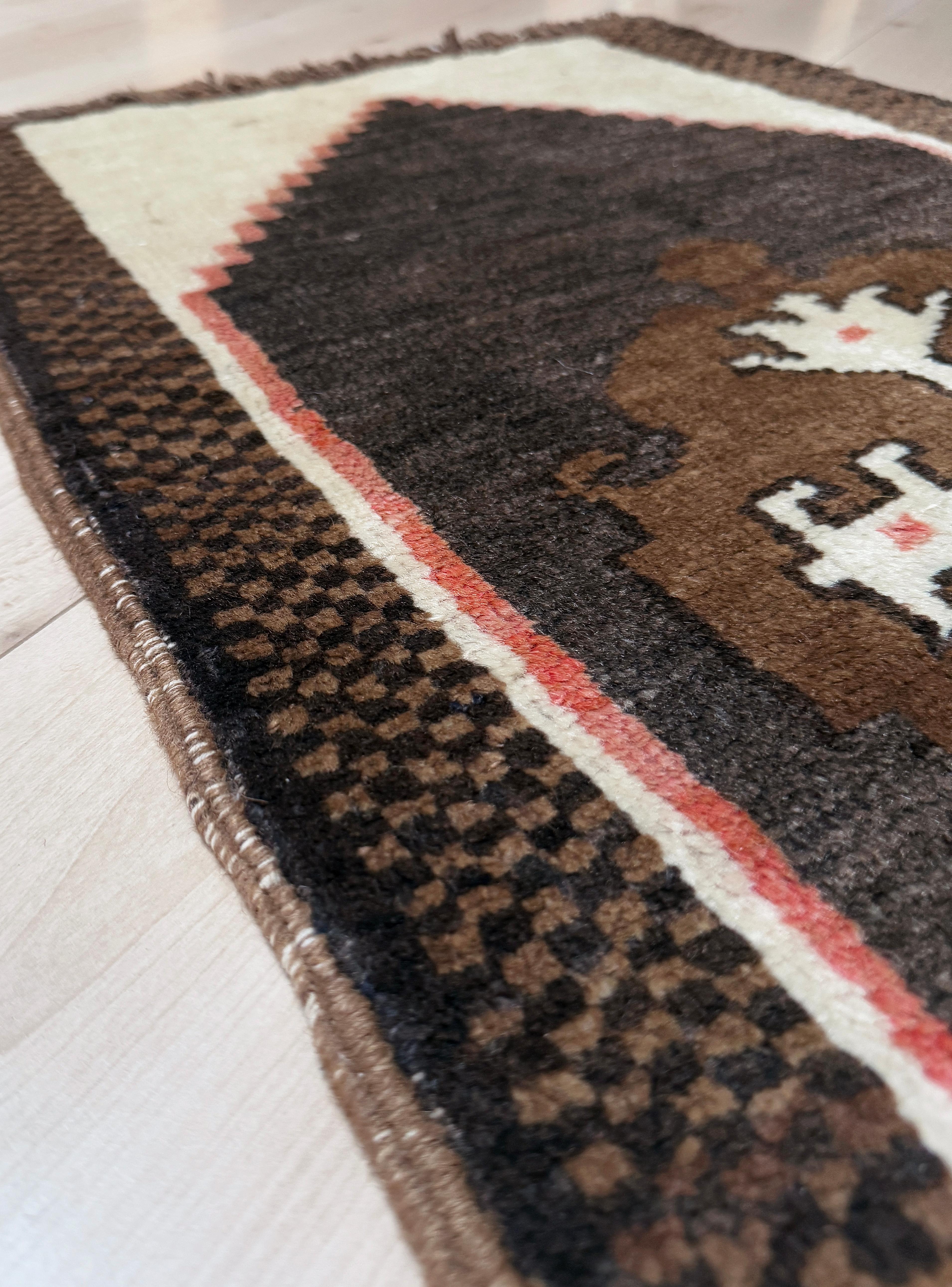 Un mini tappeto turco vintage pieno di carattere, tessuto a mano con morbida lana filata a mano e caratterizzato da un audace emblema tribale centrale incorniciato da ricche tonalità della terra. Le sue dimensioni compatte di 19 