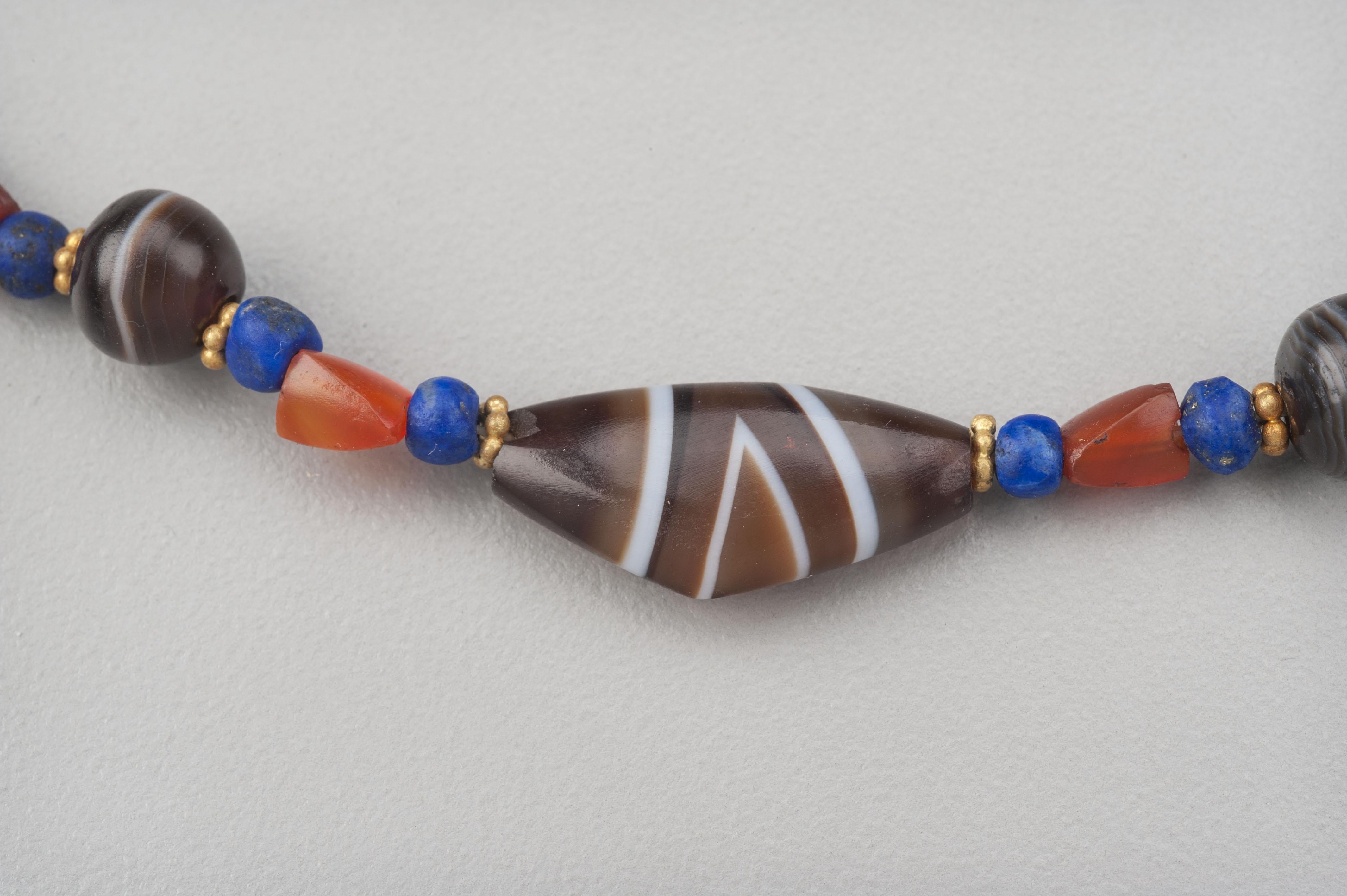 Twenty-one agate beads, nine of which are “bow” beads and twelve are round, thirteen small etched carnelian beads and forty-two lapis lazuli beads. Each of the agate beads are faced with granulated 20k gold ring beads. There are forty-two of these.