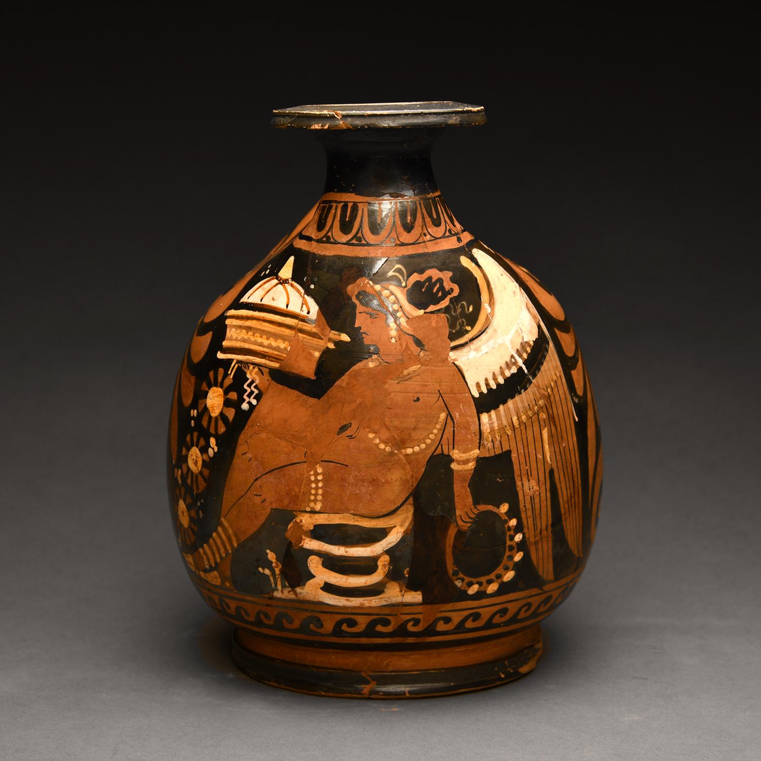 This beautifully proportioned South Italian ceramic production is an askos, or pouring vessel.  It prominently features a seated naked winged Eros wearing anklets, bracelets, a body chain, a necklace and earrings. His hair bound up, holding a laden