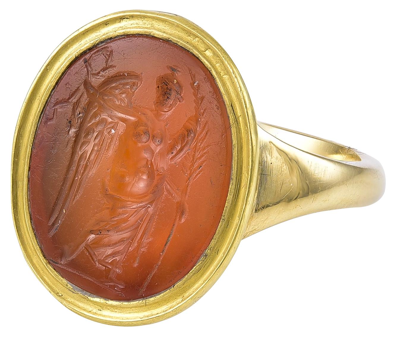 Set in an 18k Gold Ring, the Carnelian of a pleasant deep orange colour, the helmeted Nike is holding the Wreath of Victory in her right hand and in her left hand is a Palm Leaf and on her arm is draped her robe. She is bare breasted and her pose is