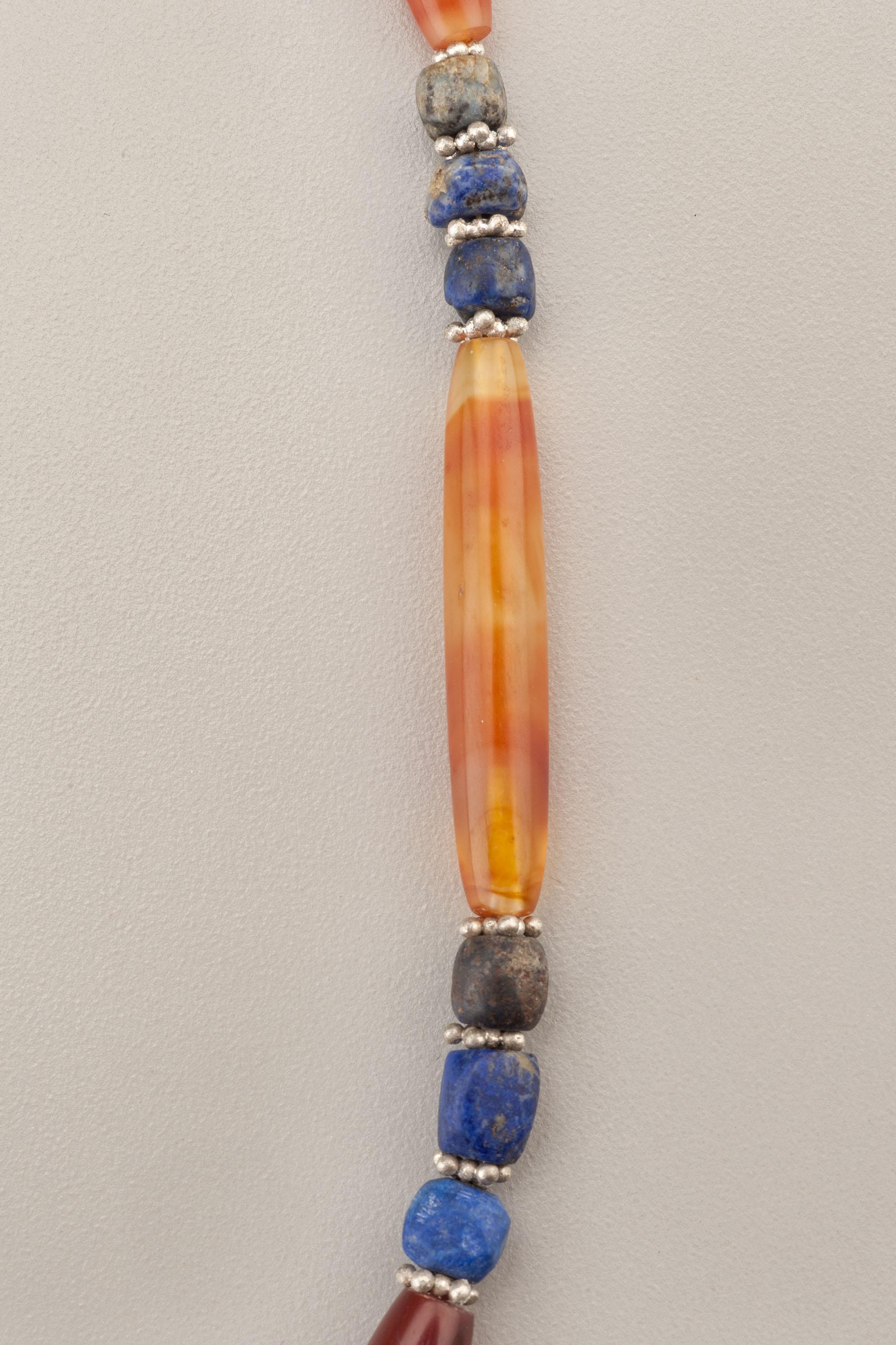 A necklace of fifteen carnelian beads, nine of which are long tapered beads and six of which are shorter barrel shaped carnelian tubes. There are fifty lapis beads in the necklace which graduate in size towards the back of the necklace. These are