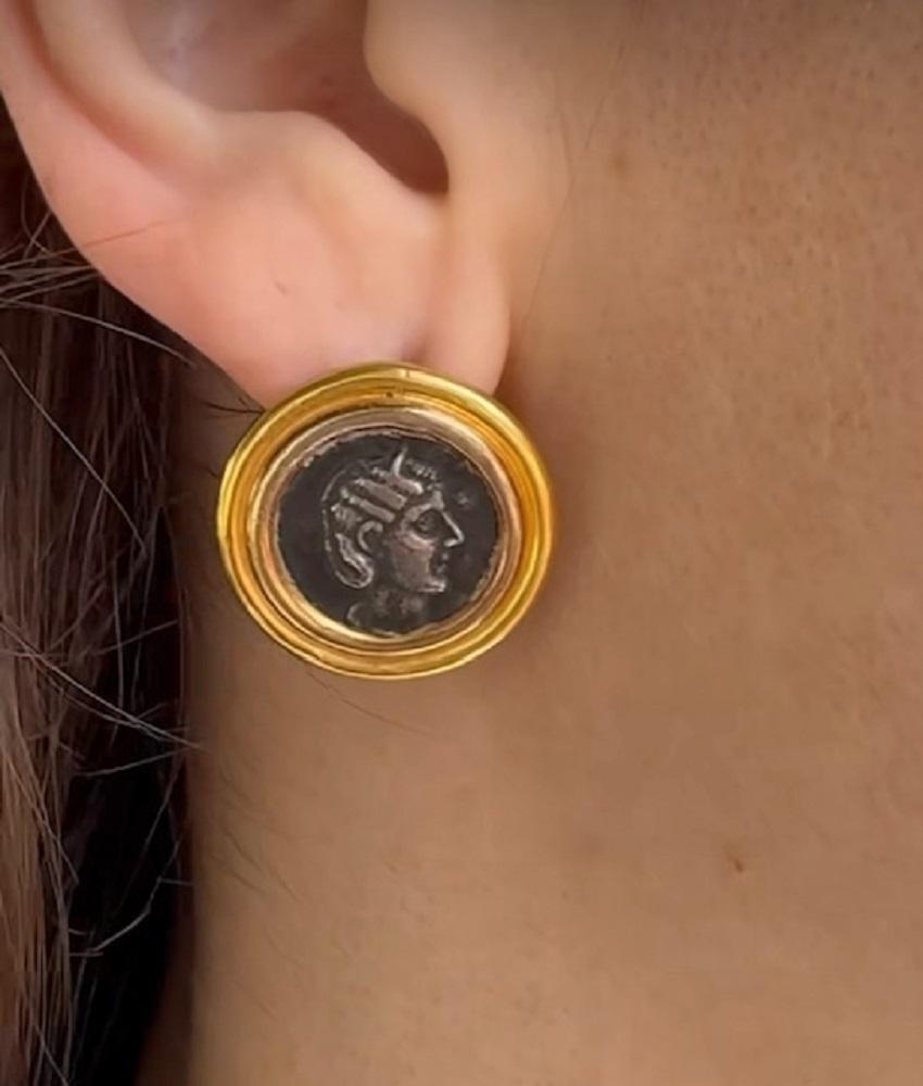 A pair of ancient coin earrings made of 22k gold.
The earrings are circular, with the silver coins set in the smooth gold frames. 
The gold creates warm and radiant contrast to the darker tones of the silver coins. Each coin showcases intricate
