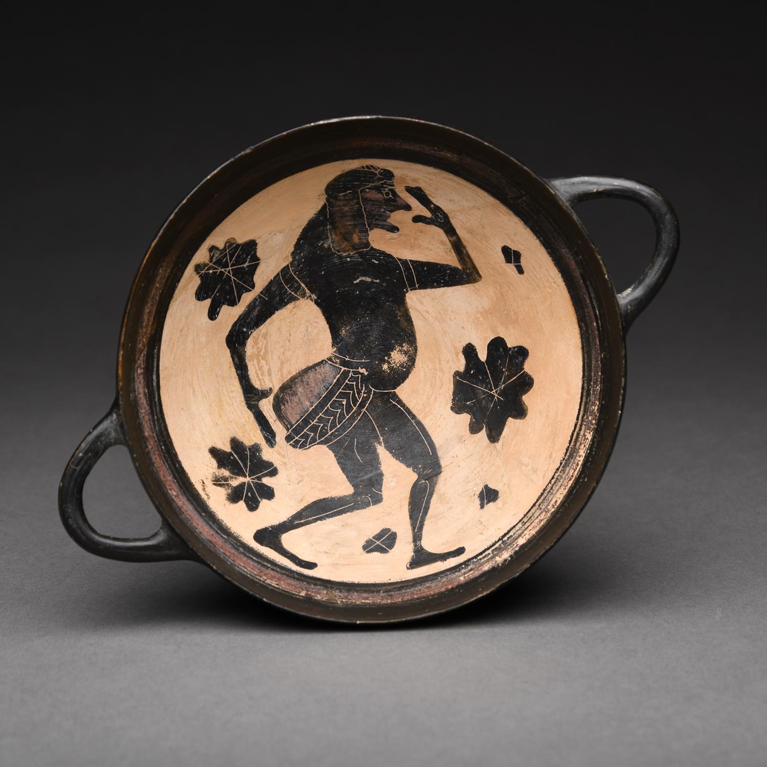 Extremely spirited, the interior of this elegant cup displays a figure of a dancing fat-bellied komast.  He wears a short padded tunic with exaggerated buttocks, and is surrounded by rosettes in the field. The black exterior features thin pointed