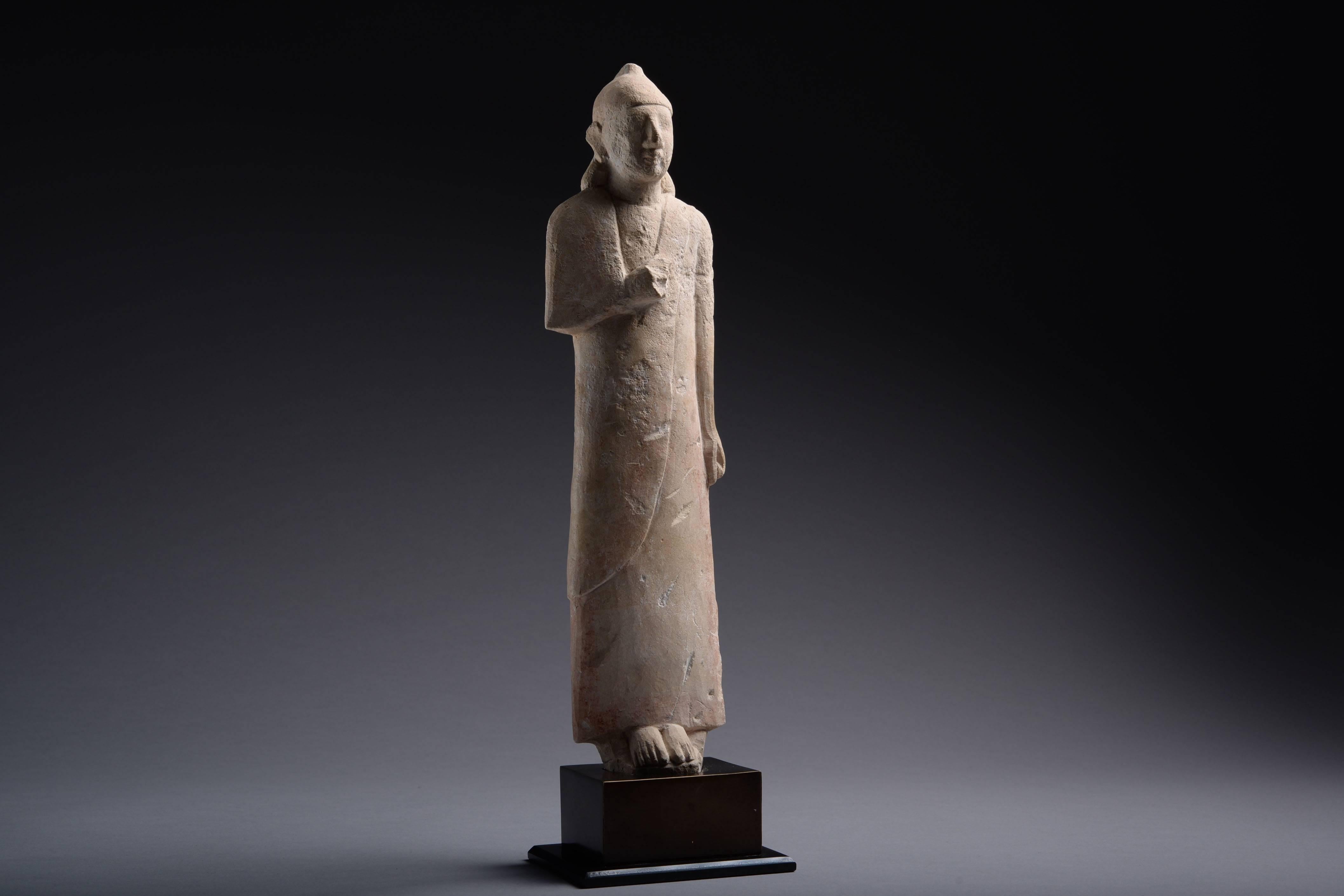 A limestone votive worshipper from Cyprus, dating to the Cypro Archaic Period, circa 600 BC. 

The male figure is shown standing tall, wearing a long tunic, his right hand folded over his chest, his left by his side, his hair pushed back over his