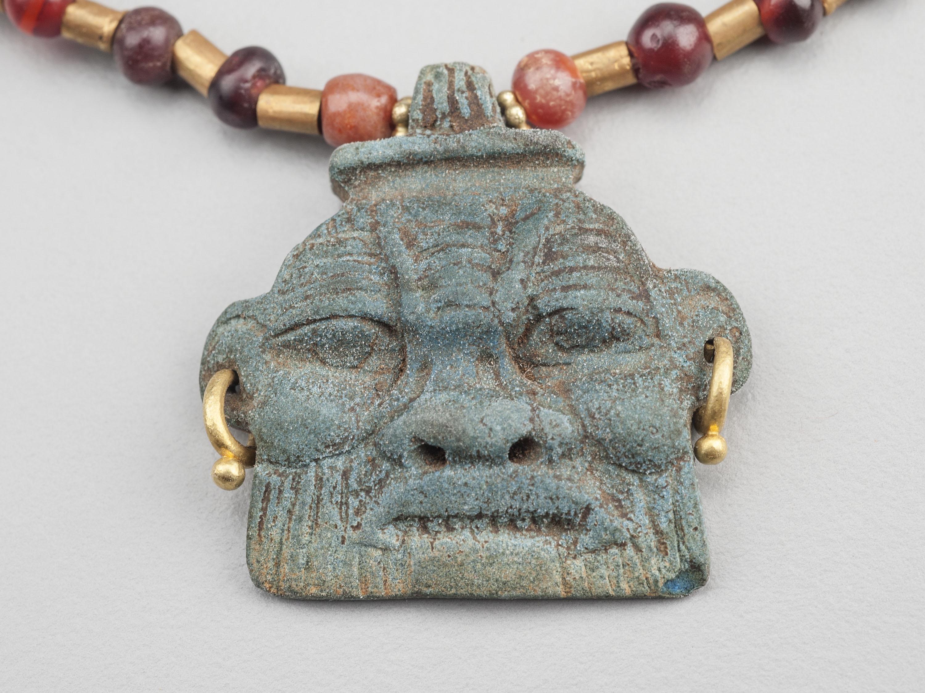 Fifty four garnet beads alternating with fifty eight 20k gold tube beads with two carnelian bi cone barrel beads midway back on each side of the necklace; a faience pendant depicting the ancient Egyptian deity Bes hangs from the center of the