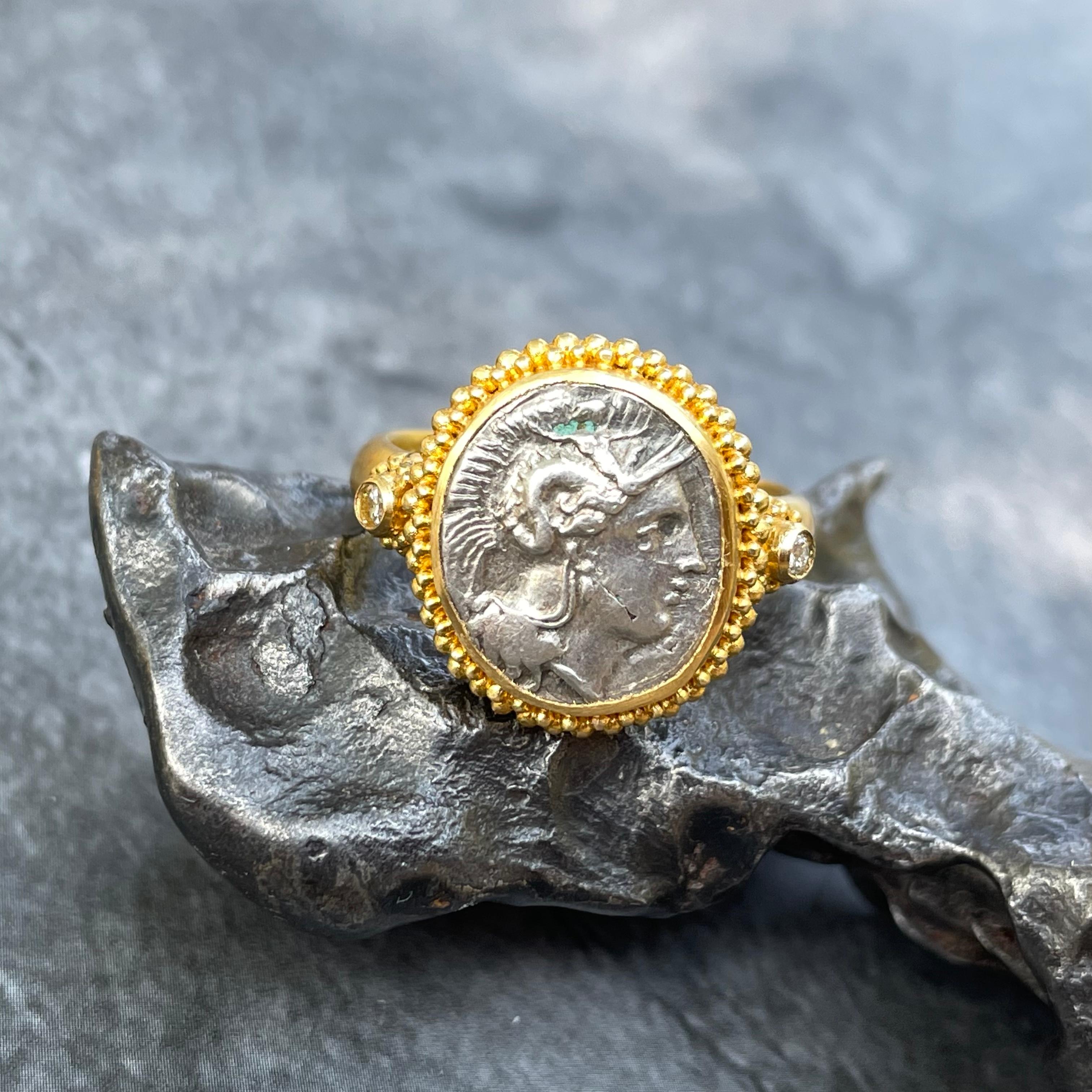 A authentic ancient coin with classic image of the goddess Athena wearing a crested helmet is displayed between two 1.8 MM diamonds and surrounded by stacked large and small hand applied granulation in this Steven Battelle high carat gold creation. 