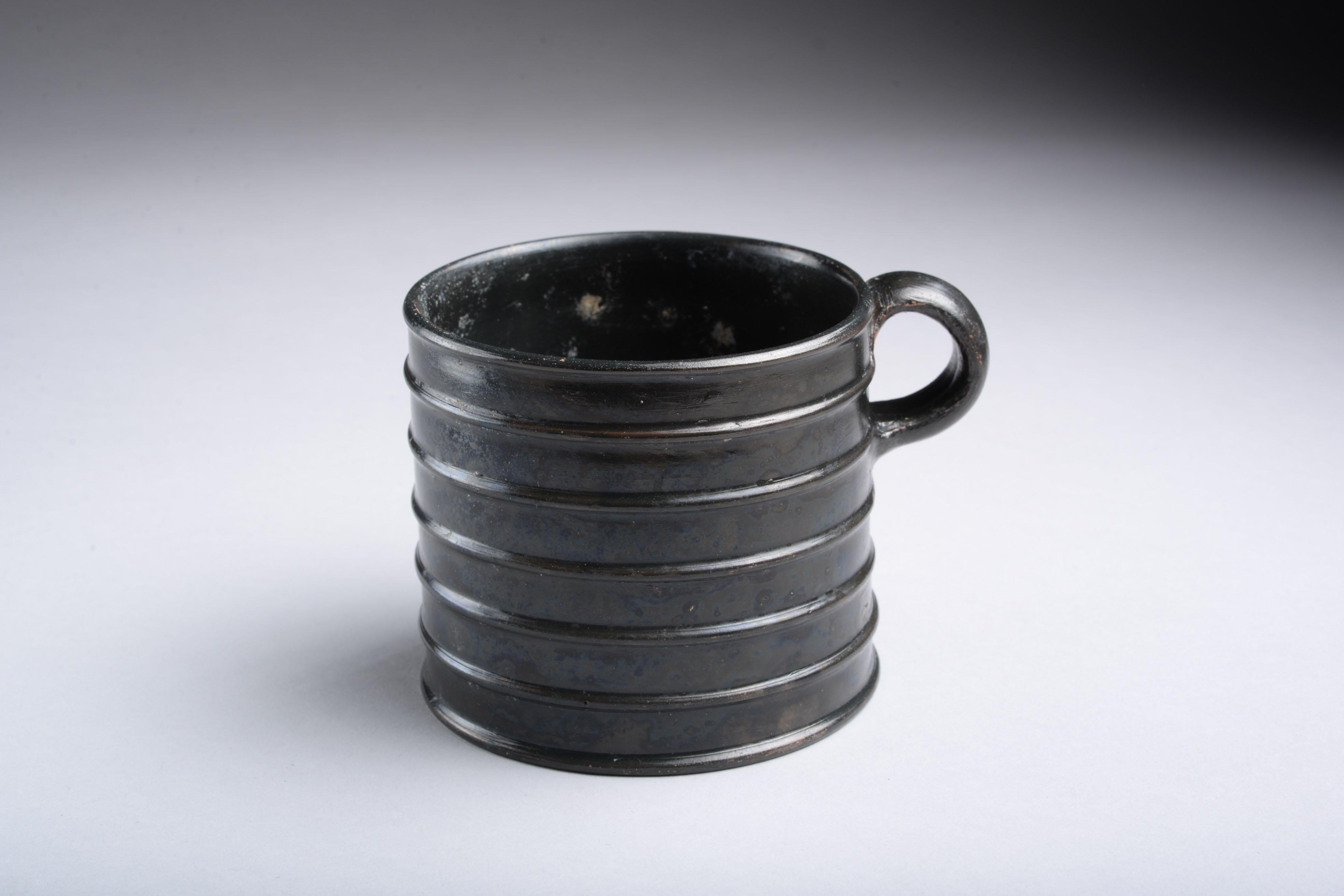 Greek black-glaze ribbed mug
Athens, circa 475-425 B.C.
terracotta.

The mug is of a straight-sided, cylindrical form with seven horizontal ribs and an applied ring handle just below the lip. The base is flat with a slightly raised foot running