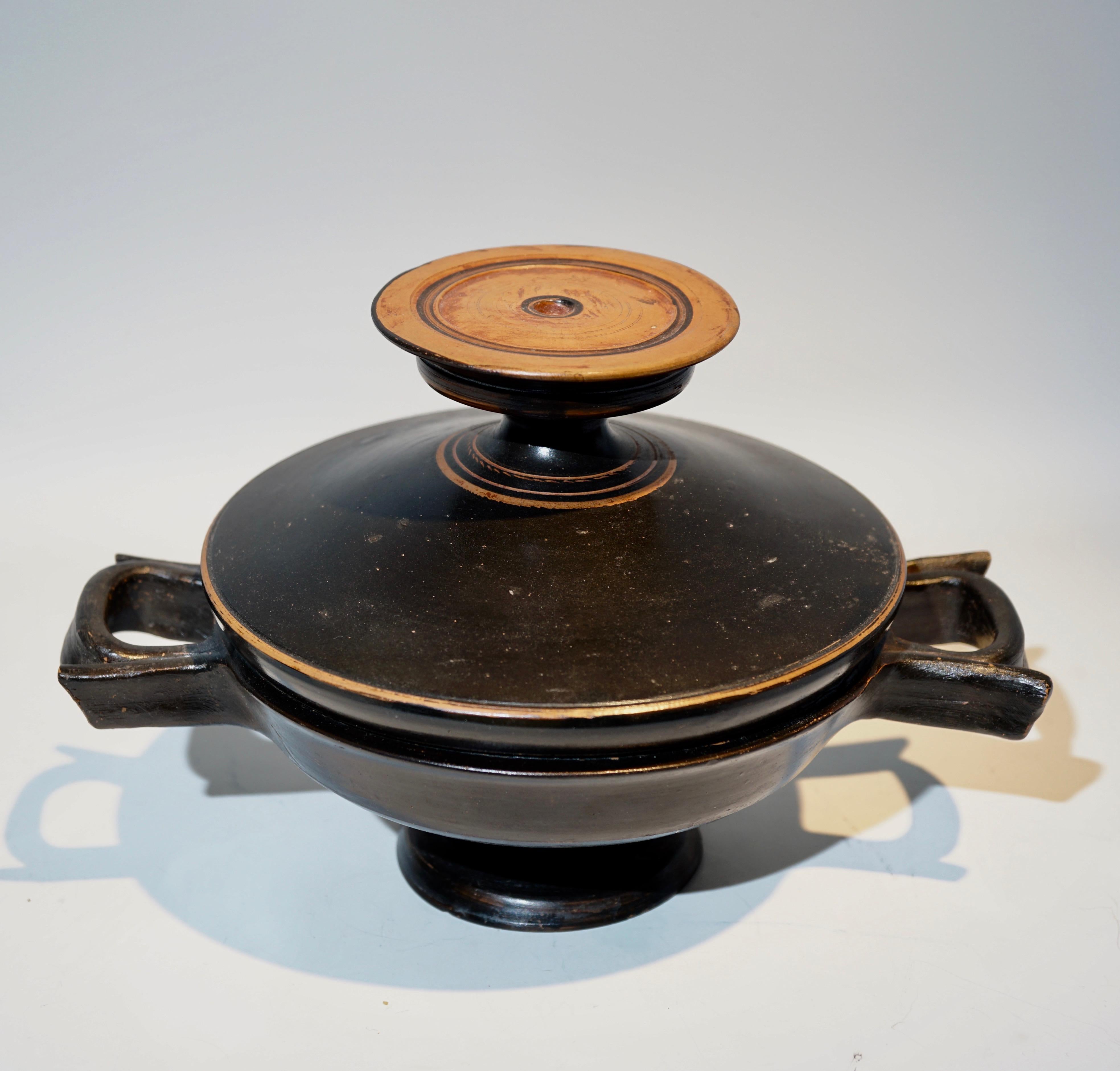 Attic sizeable black glazed lidded lekanis with ribbon-like handles projecting from the sides, resting on a broad foot. The lid is topped with a large disc-like knob with slightly raised edges that is sitting on a short stem. The lid is decorated