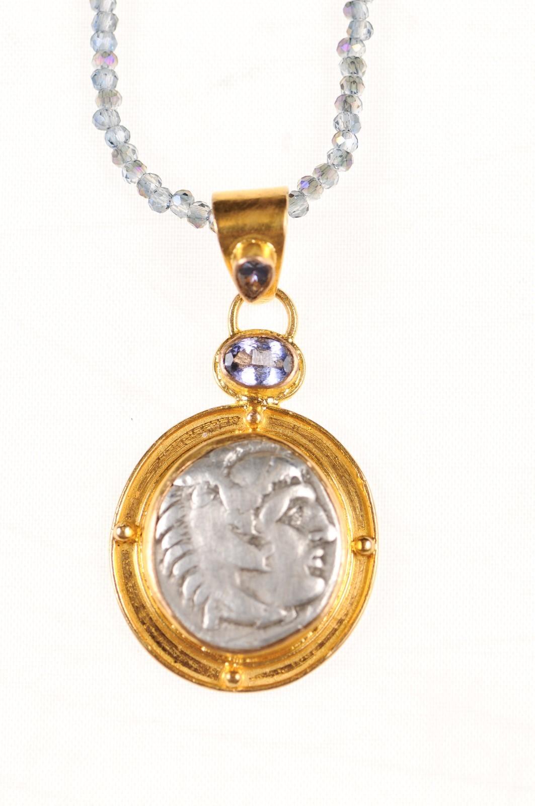 An Authentic Greek (Macedonian) Alexander the Great silver Drachm coin (circa 336 - 323 BC) set in a 22k gold bezel and bail adorned with tanzanite stones. The obverse, or front, side of this coin features Heracles. On the reverse side, Zeus seated