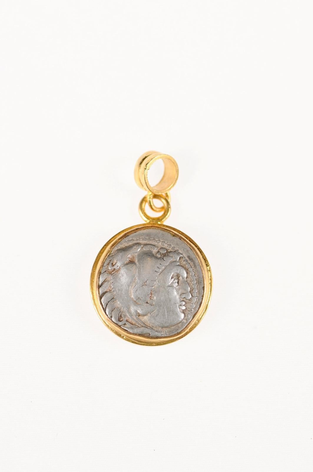 An authentic Greek, Alexander the Great, AR Drachm coin (circa 3rd century BC), set in a round 22k gold bezel with 22k gold bail. The obverse, or front side of this coin features the head of Herakles wearing a lion skin headdress. On the reverse