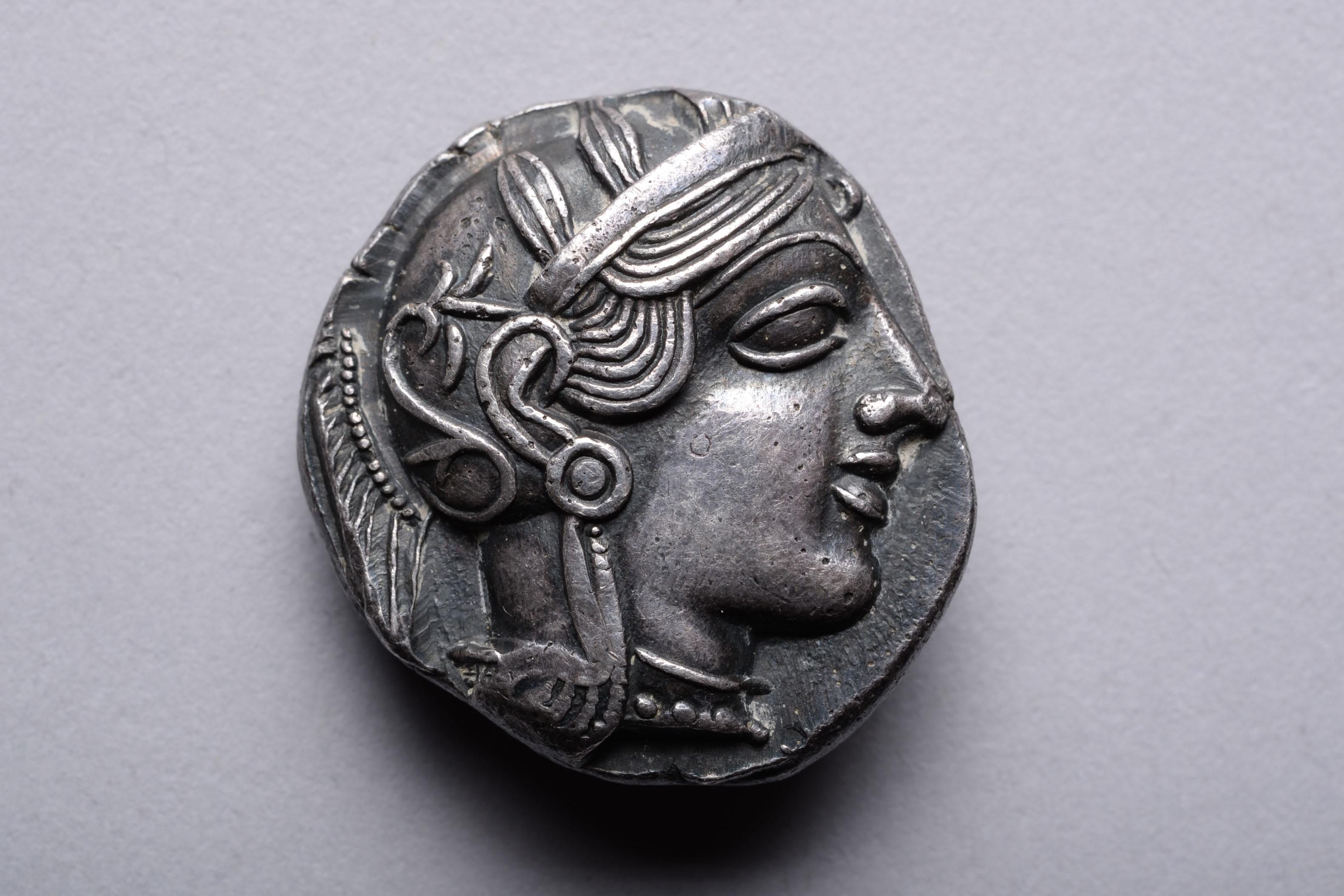 Silberne Tetradrachme aus Athen, ca. 454-404 v. Chr.

Die Vorderseite der Münze zeigt Athena, die Schutzgöttin von Athen. Die Göttin trägt eine Perlenkette und einen Helm mit Kamm, der mit Olivenblättern und einer Blumenrolle verziert ist, ihr