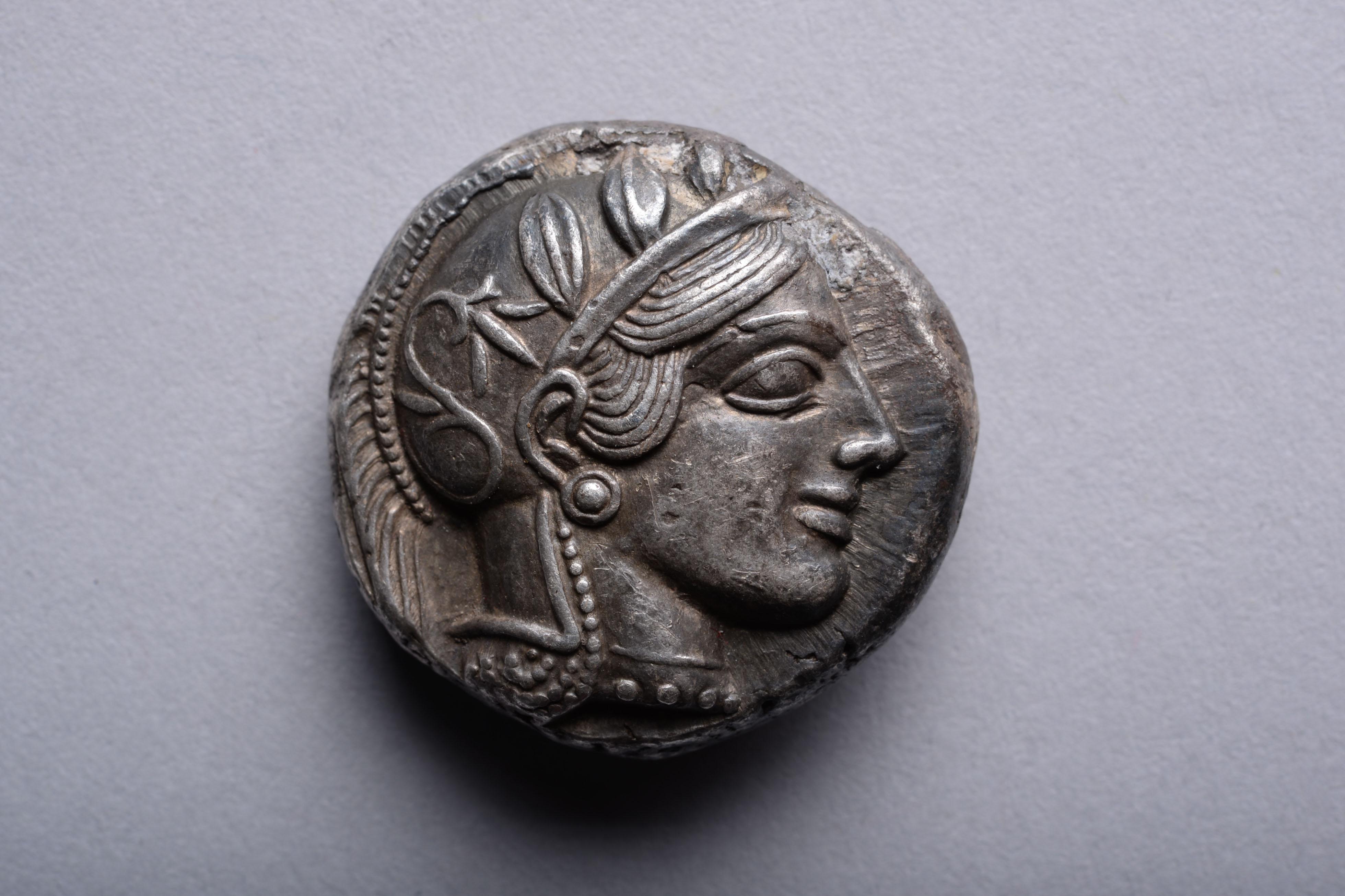 A silver tetradrachm from Athens, circa 454-404 BC.

The front of the coin with Athena, patron deity of Athens. The goddess wears a beaded necklace and crested helmet, decorated with olive leaves and a floral scroll, her hair swept across her