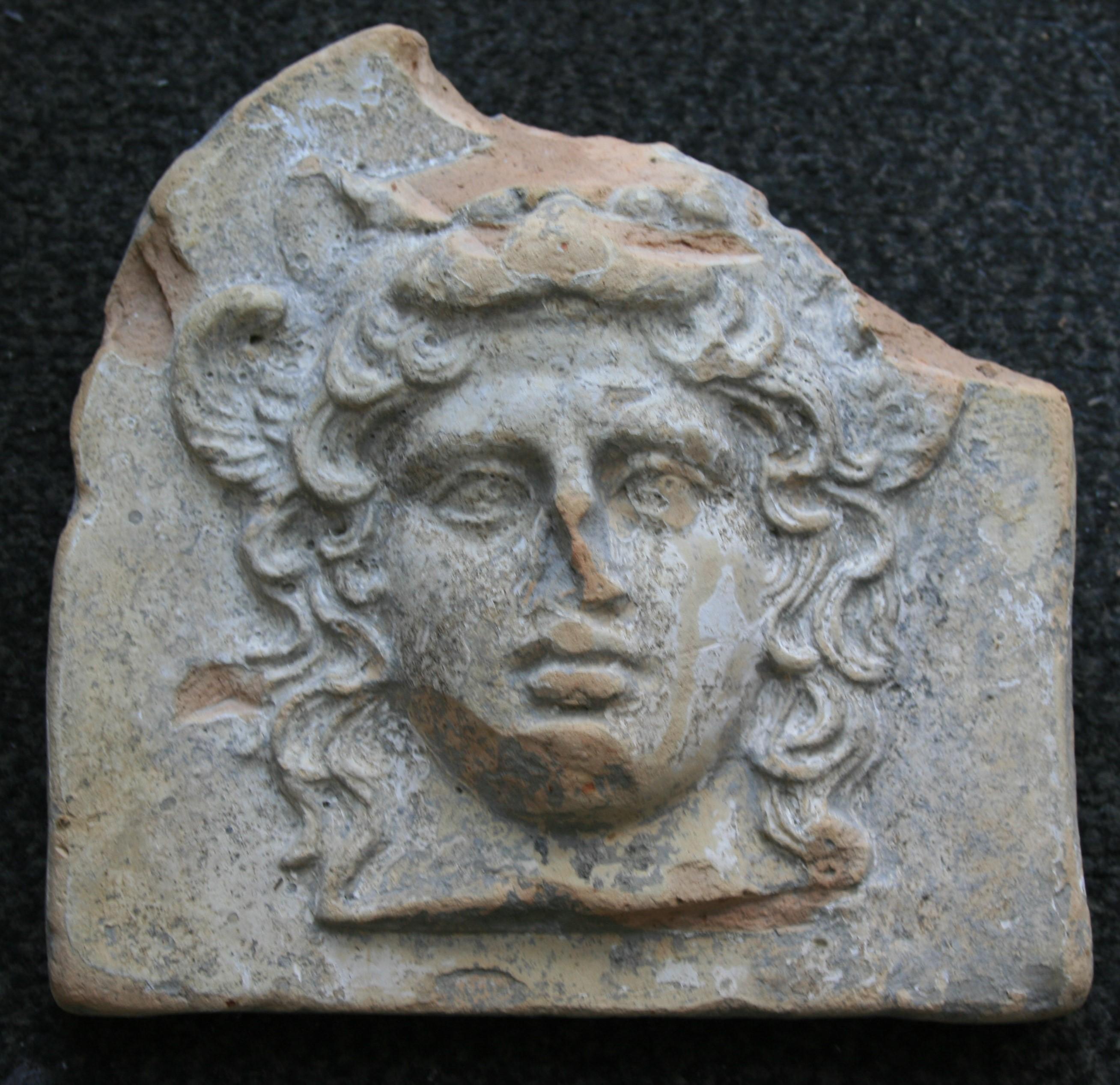 Altgriechisches Terrakotta-Antefix, verziert mit dem weiblichen Kopf der Göttin Artemis Bendis.
Westgriechisch, Italien, Tarent, frühes 4. Jahrhundert v. Chr.
Zwei kleine gefiederte Flügel, die das Gesicht der Göttin flankieren, das gewellte