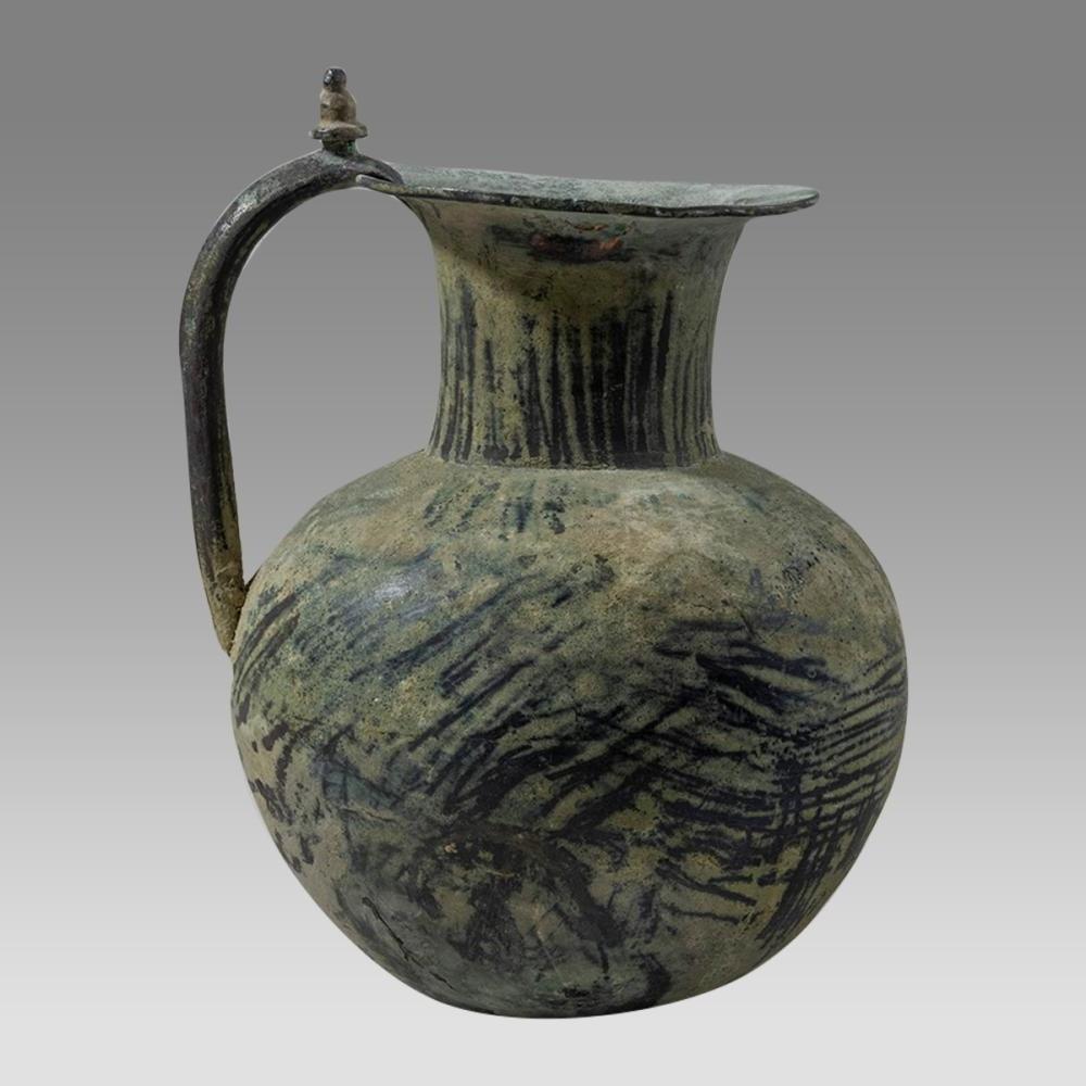 A rare and large Luristan bronze ewer, dating to circa 1000 BC. The vessel is finely cast with a rounded body, tall flaring neck, and a sharply everted rim, joined by an arched bronze handle terminating in a small knop finial. Traces of painted